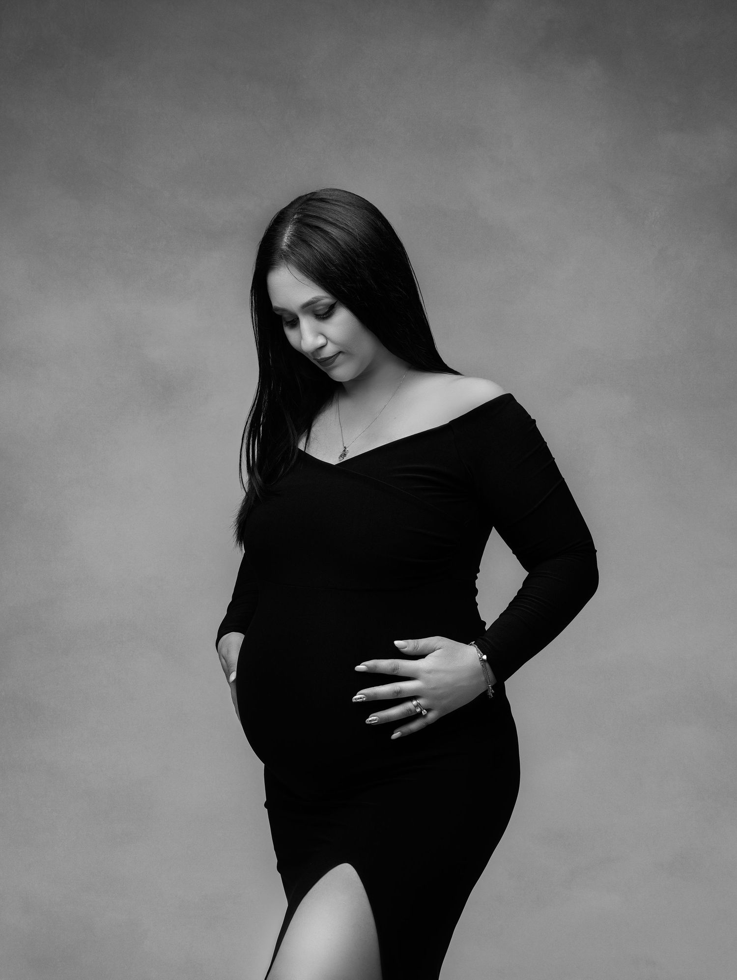 Black and white fine art portrait of an expectant mother in long-sleeved dress posing elegantly against textured backdrop.