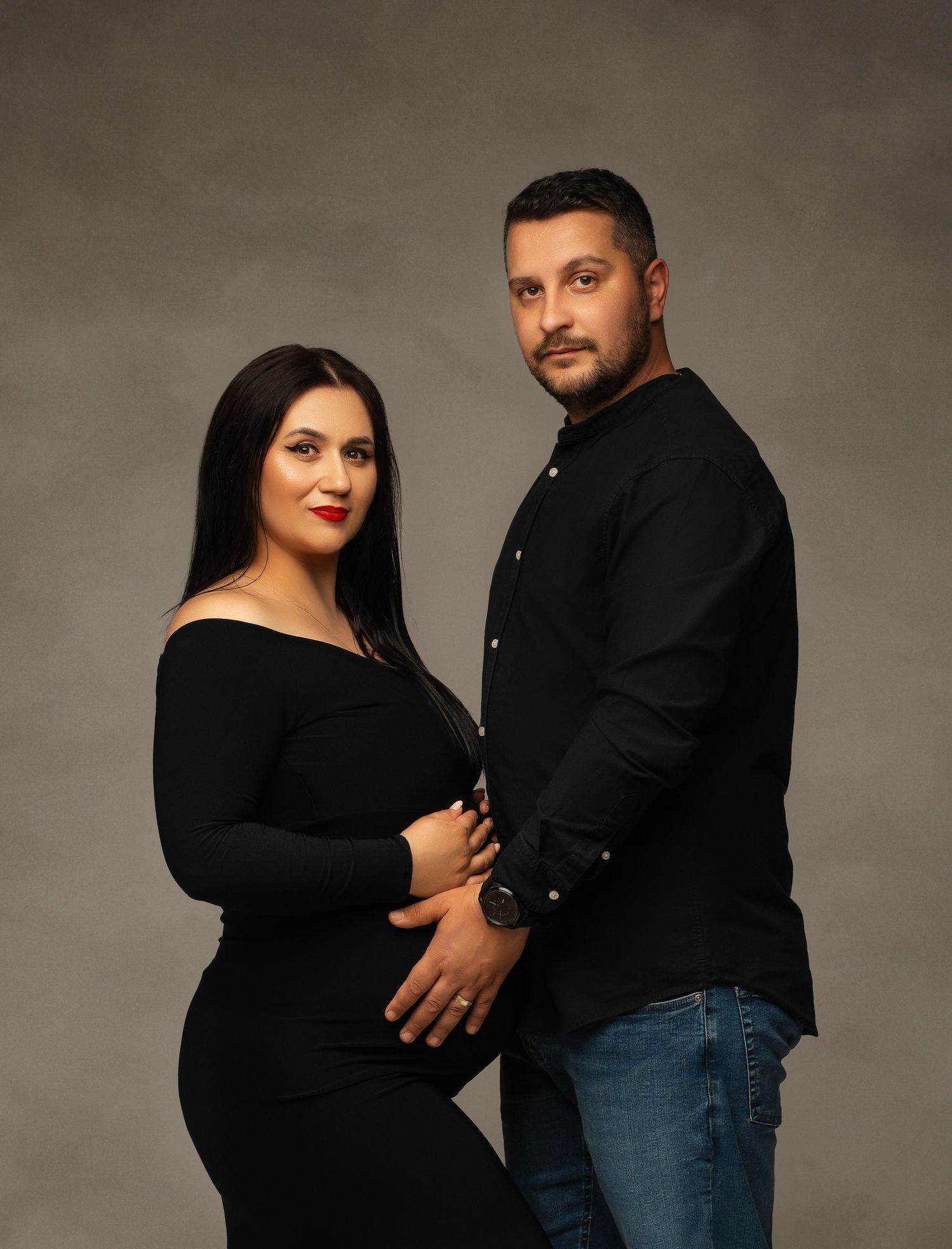 Intimate studio portrait of an expectant couple wearing black tops against a dark gray backdrop.