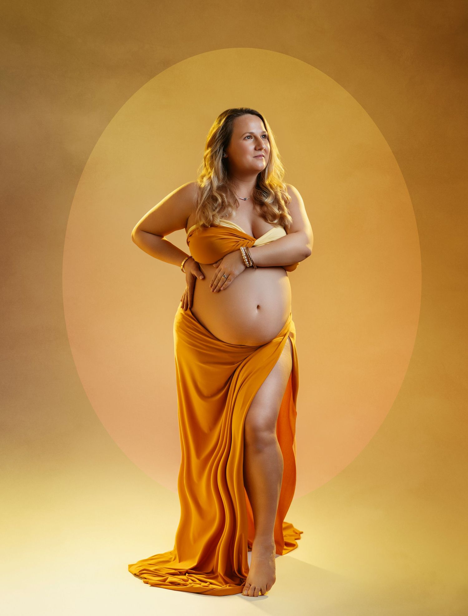 Maternity portrait of an expectant mother wearing a flowing golden dress against a warm-toned backdrop.