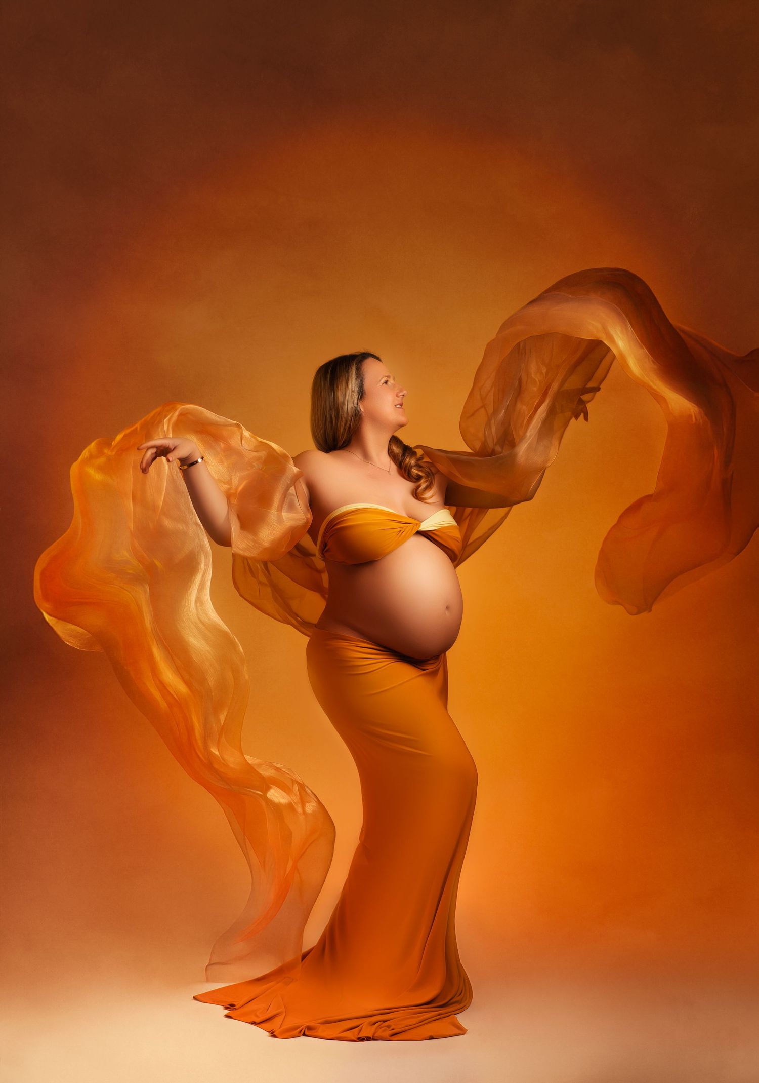 Ethereal maternity portrait in warm orange tones with flowing fabric creating dramatic movement against amber backdrop.