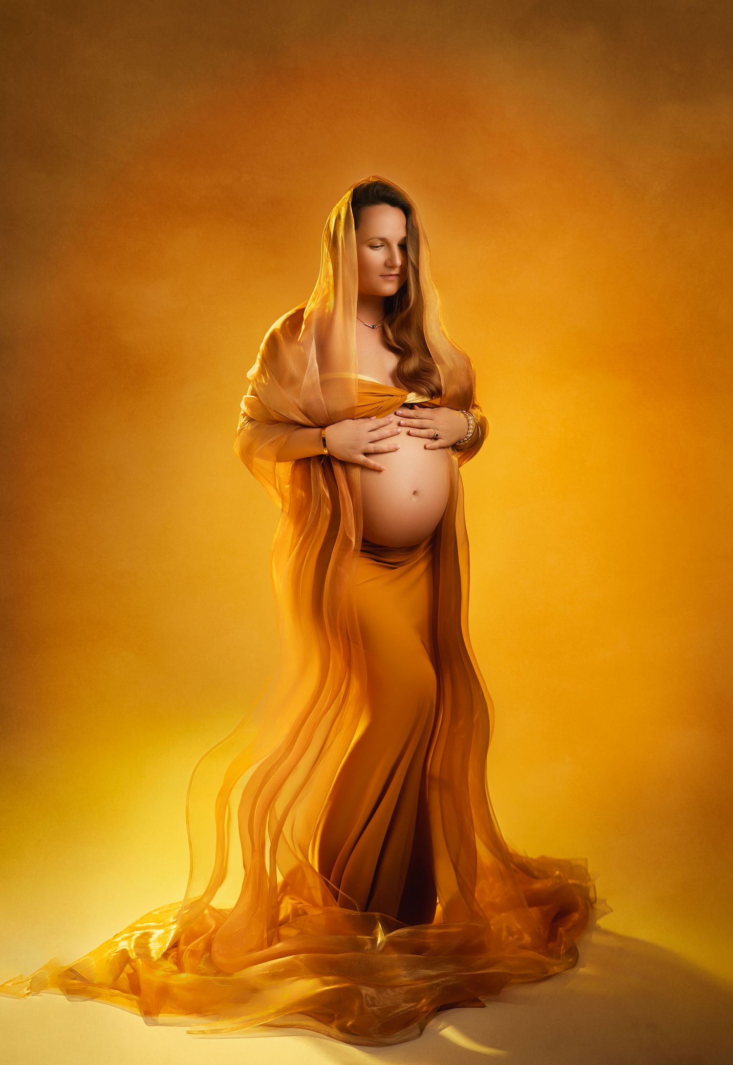 Maternity portrait showing a pregnant figure draped in flowing golden fabric against a warm glowing orange backdrop.