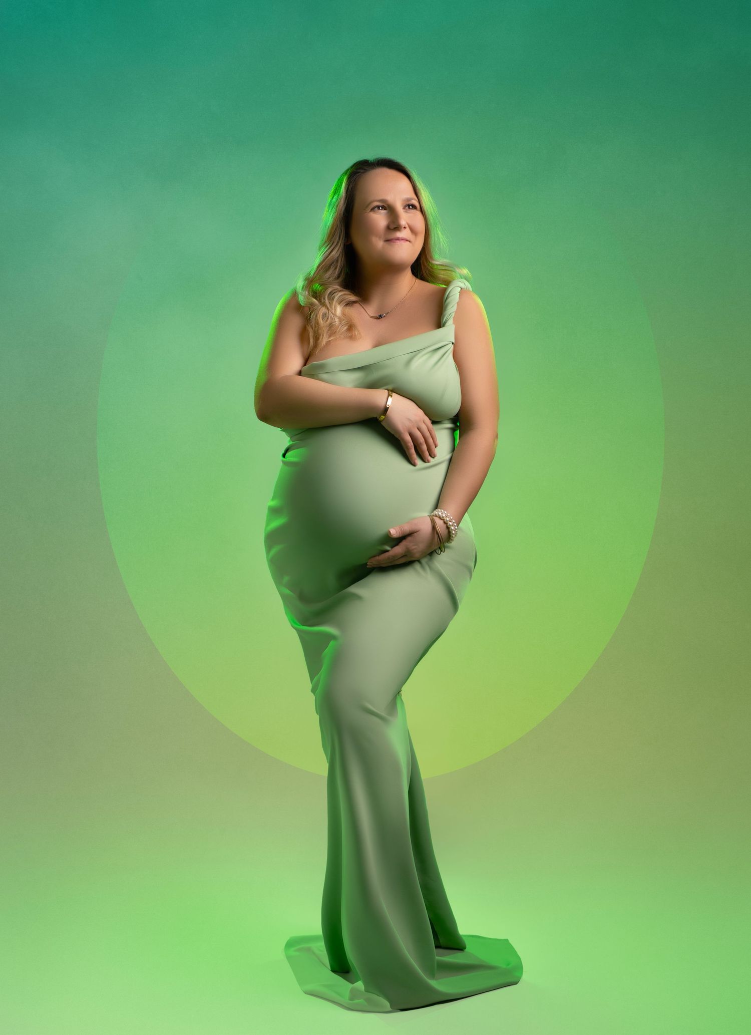 Maternity portrait against green backdrop shows figure in flowing light green gown posing elegantly in studio lighting.