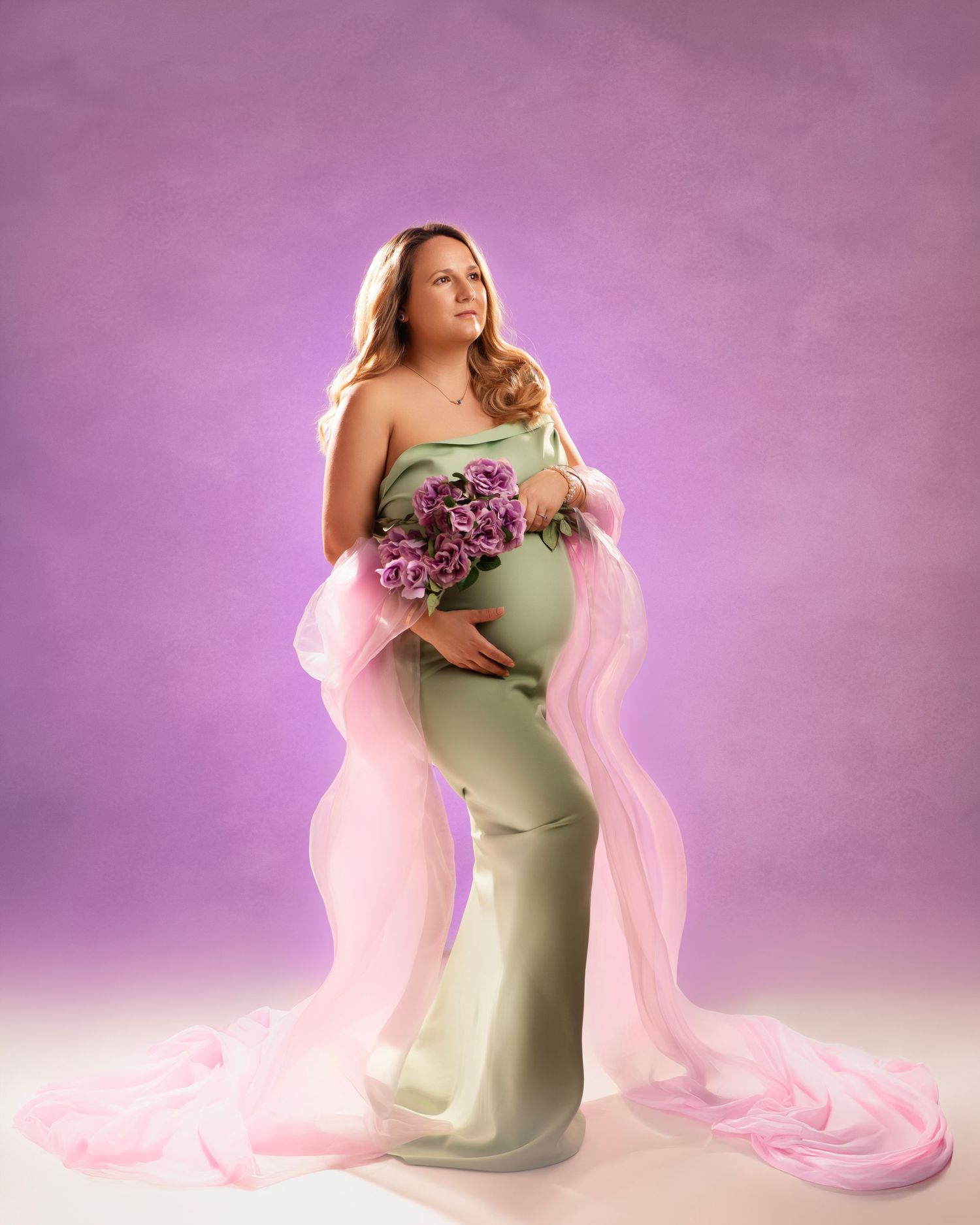 Elegant maternity portrait features a flowing pink fabric and sage green dress with purple floral bouquet against purple backdrop.