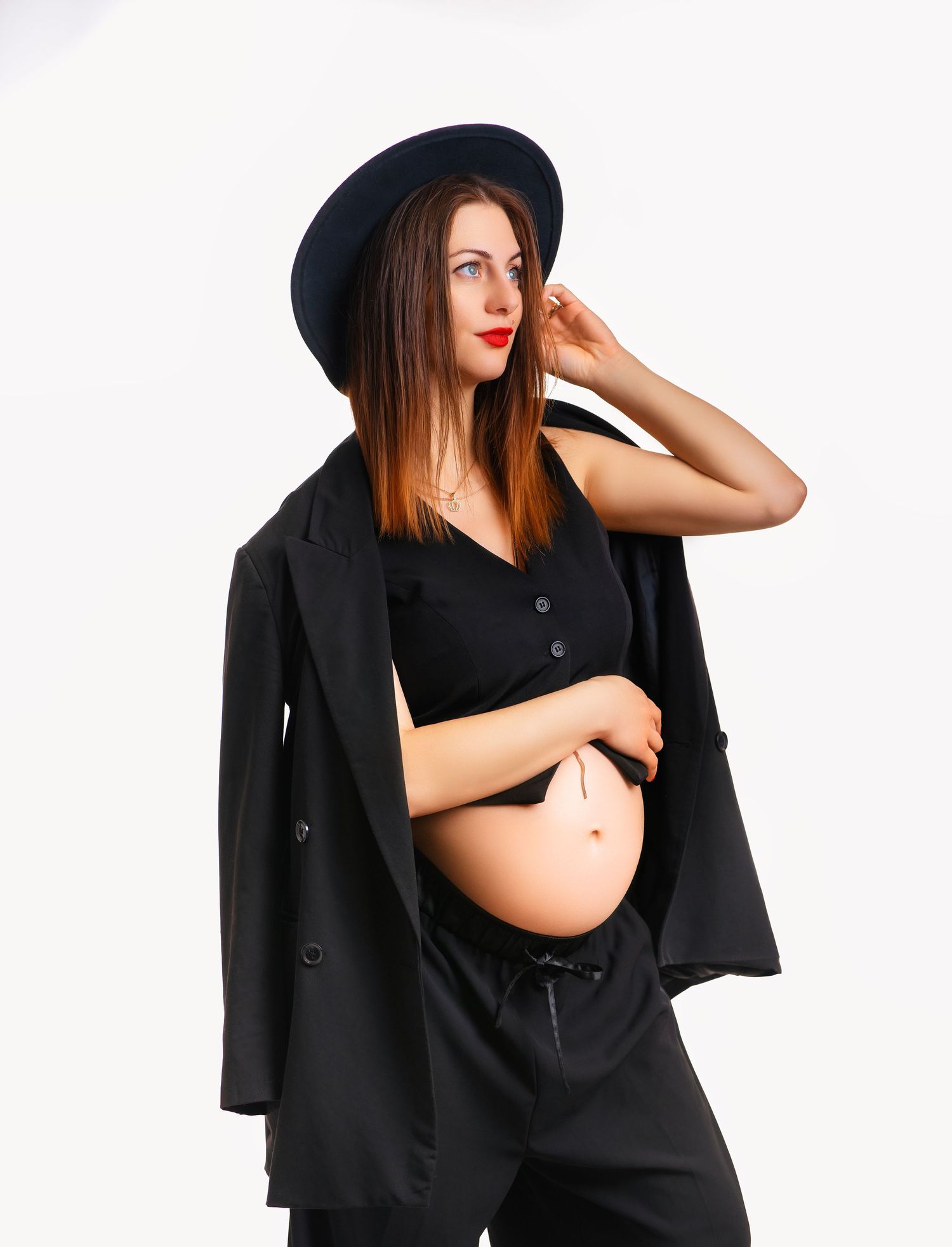 A pregnant person in a black outfit and hat poses against white background in a stylish fashion photoshoot.
