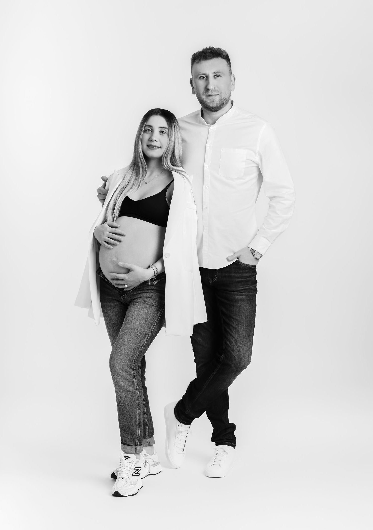 Black and white maternity photo of a couple posing together in casual attire against white background.