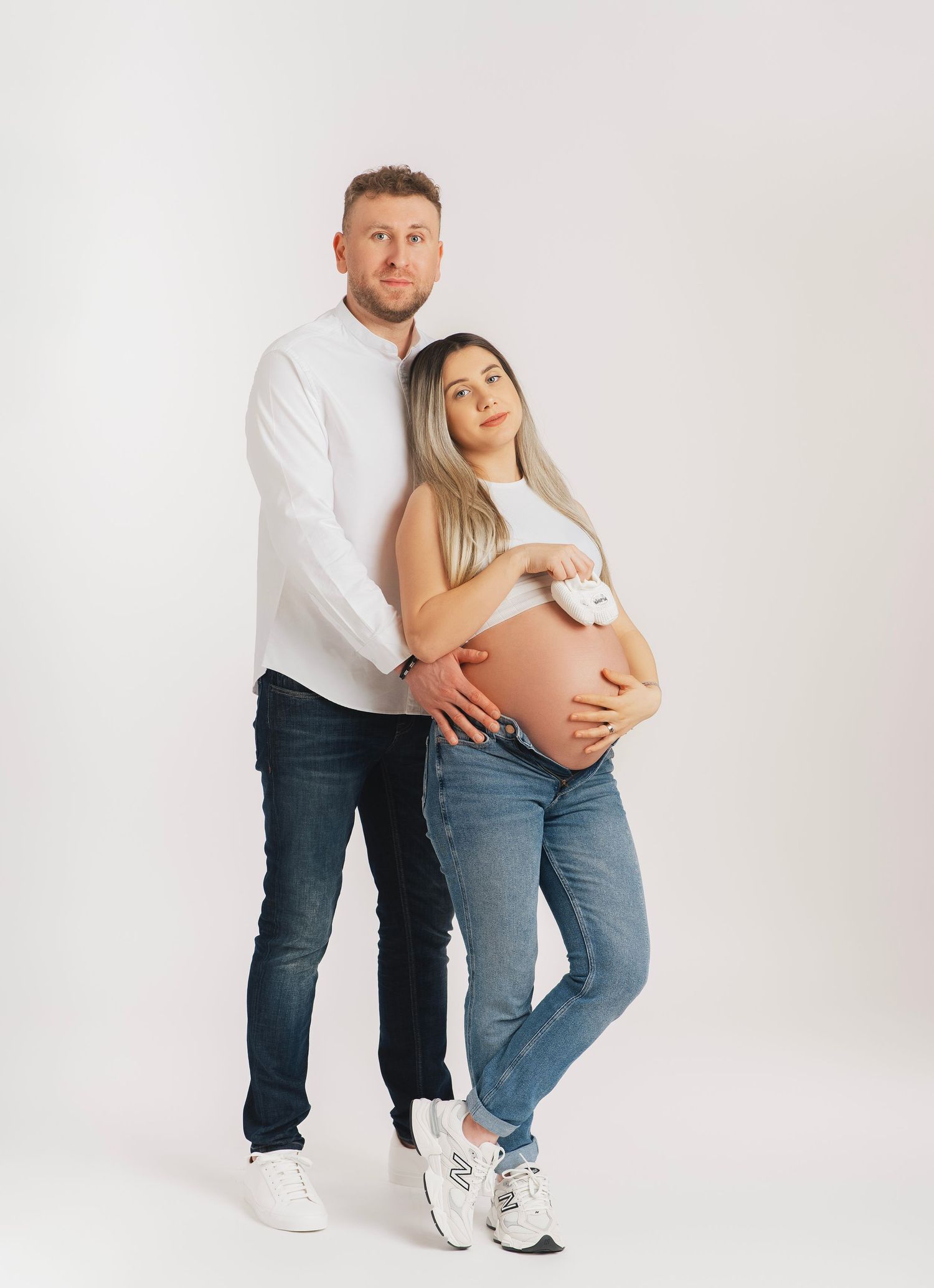 A couple poses together for a maternity photo against a white backdrop, with the woman wearing jeans and showing her baby bump.