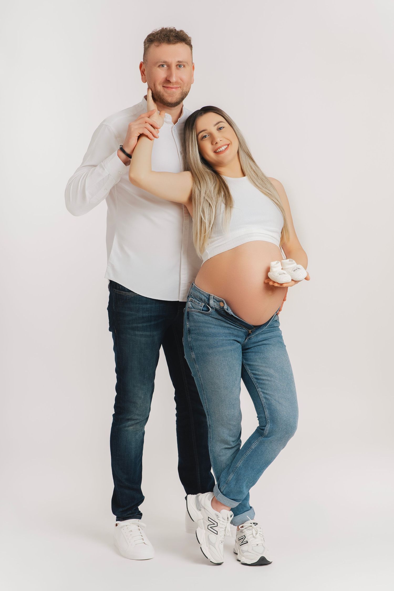 Expectant couple in casual clothing poses together against white background, showing off pregnant belly.