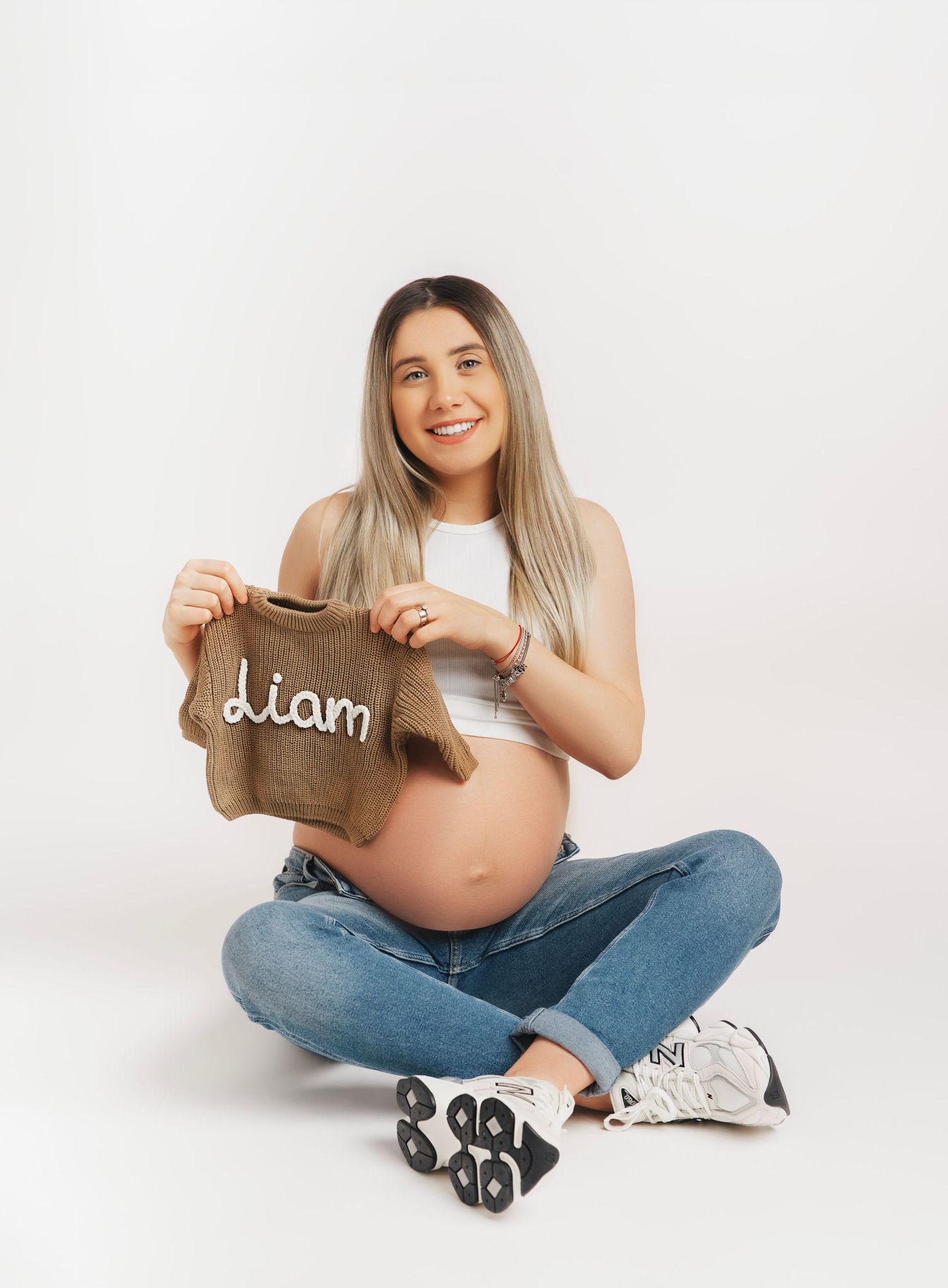 Expectant mother in casual attire sits cross-legged holding a small brown garment against white background.