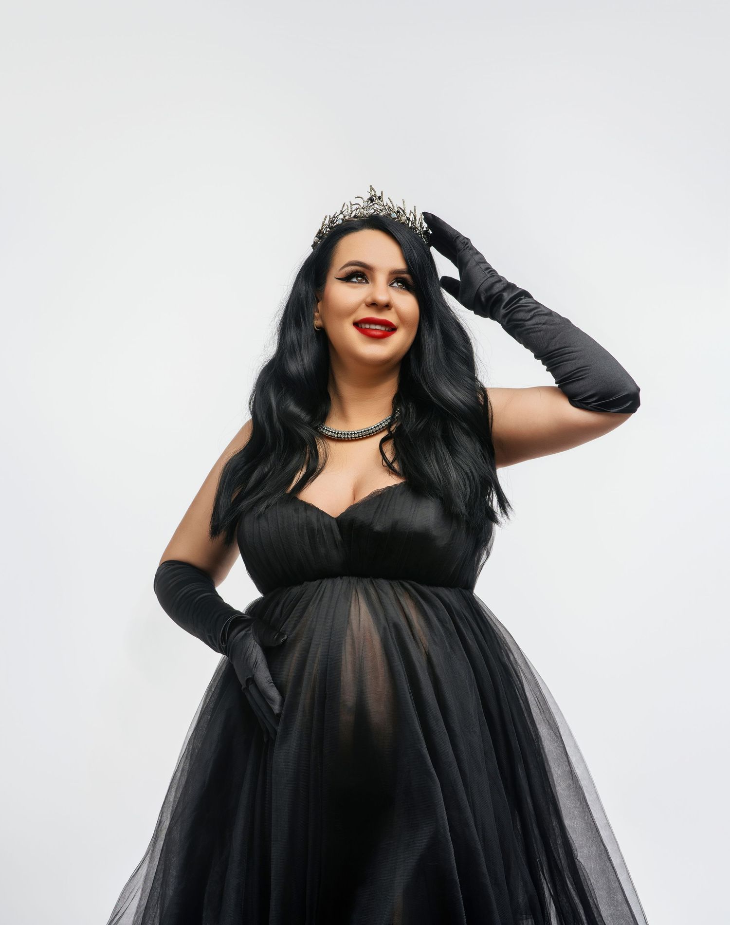 Model in elegant black tulle gown and long gloves poses against white background, styled with tiara and red lipstick.