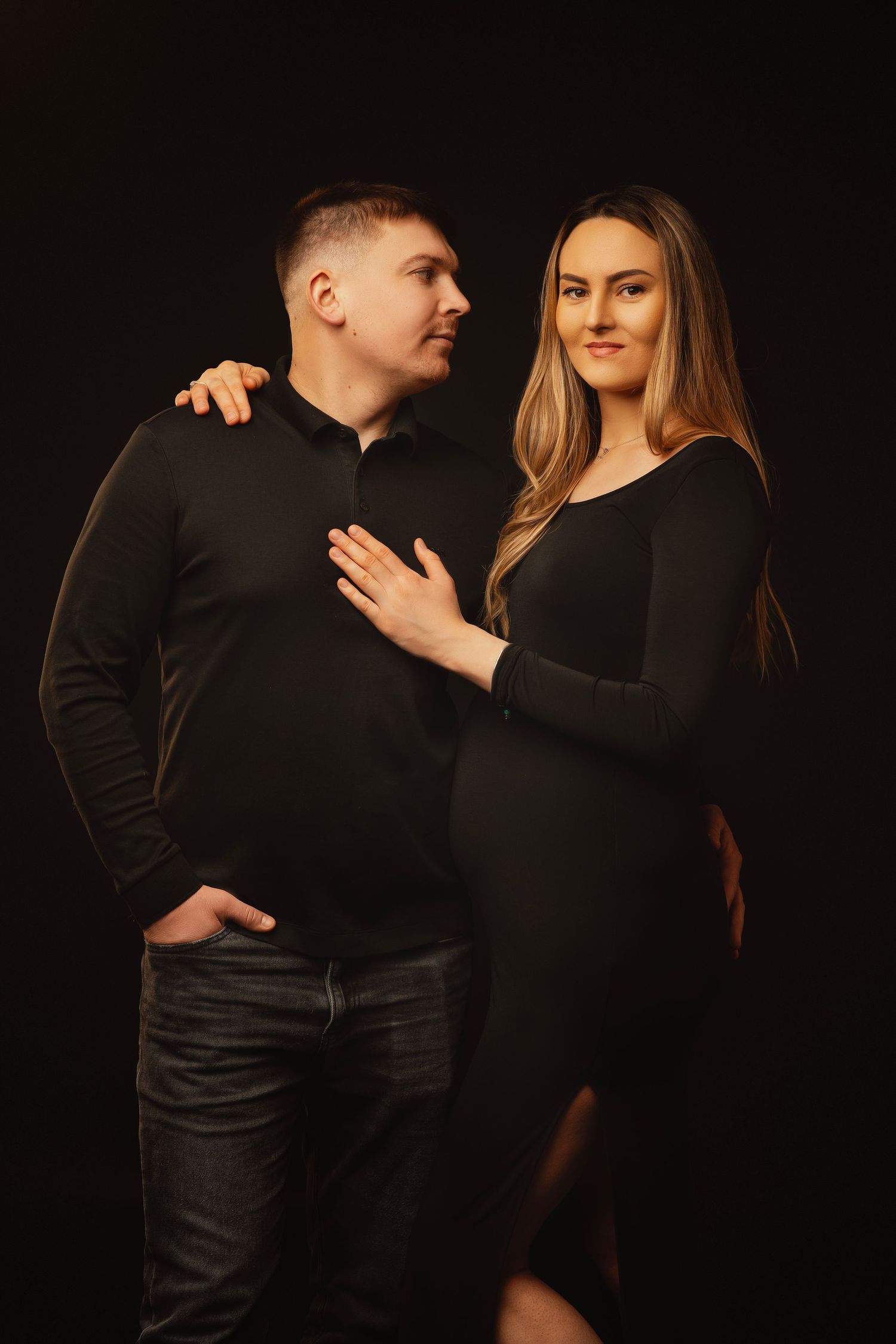A couple dressed in matching black outfits pose together against a dark background in an elegant portrait.