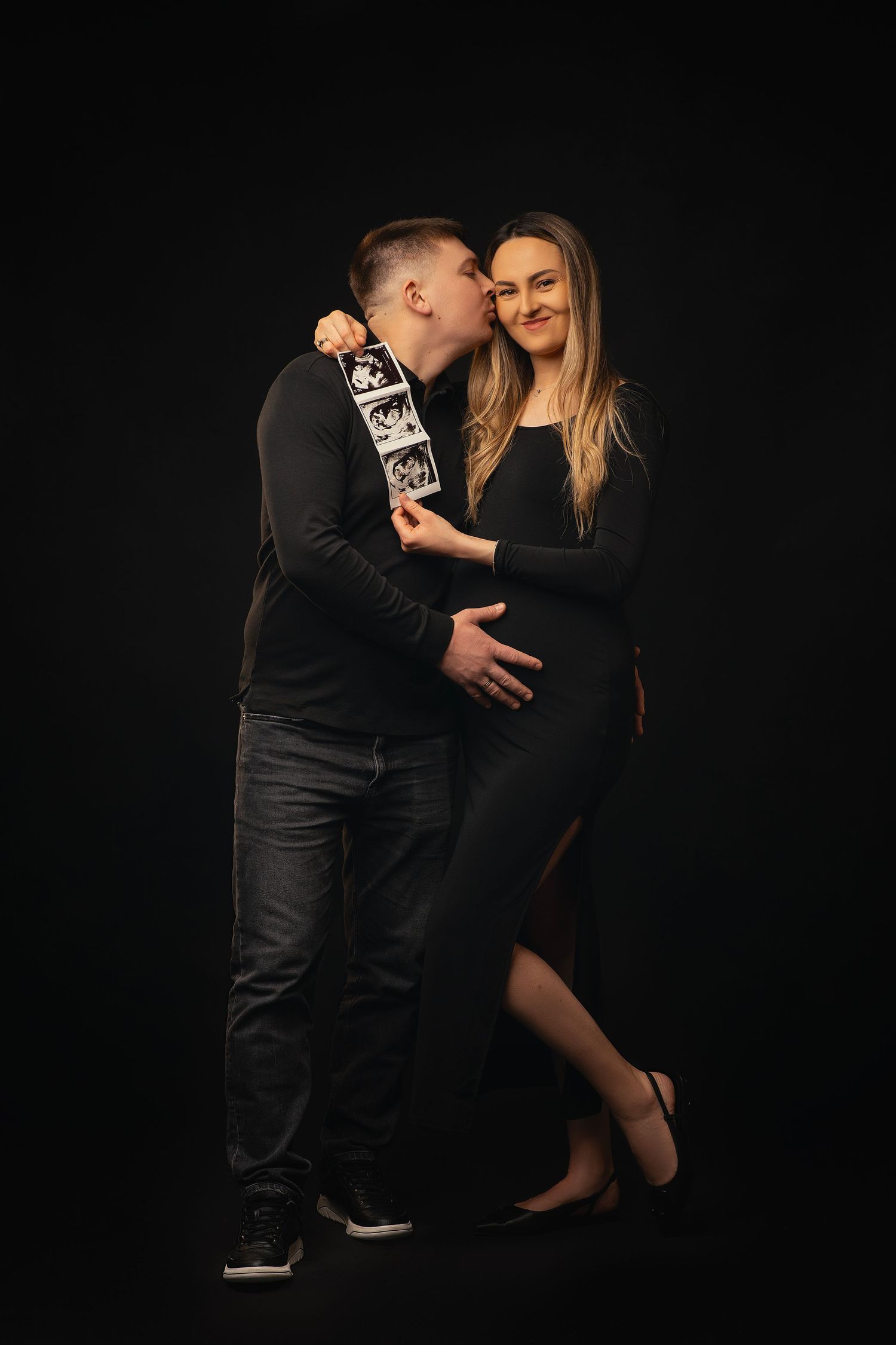 A couple in black attire poses intimately against a dark background during a romantic photoshoot.