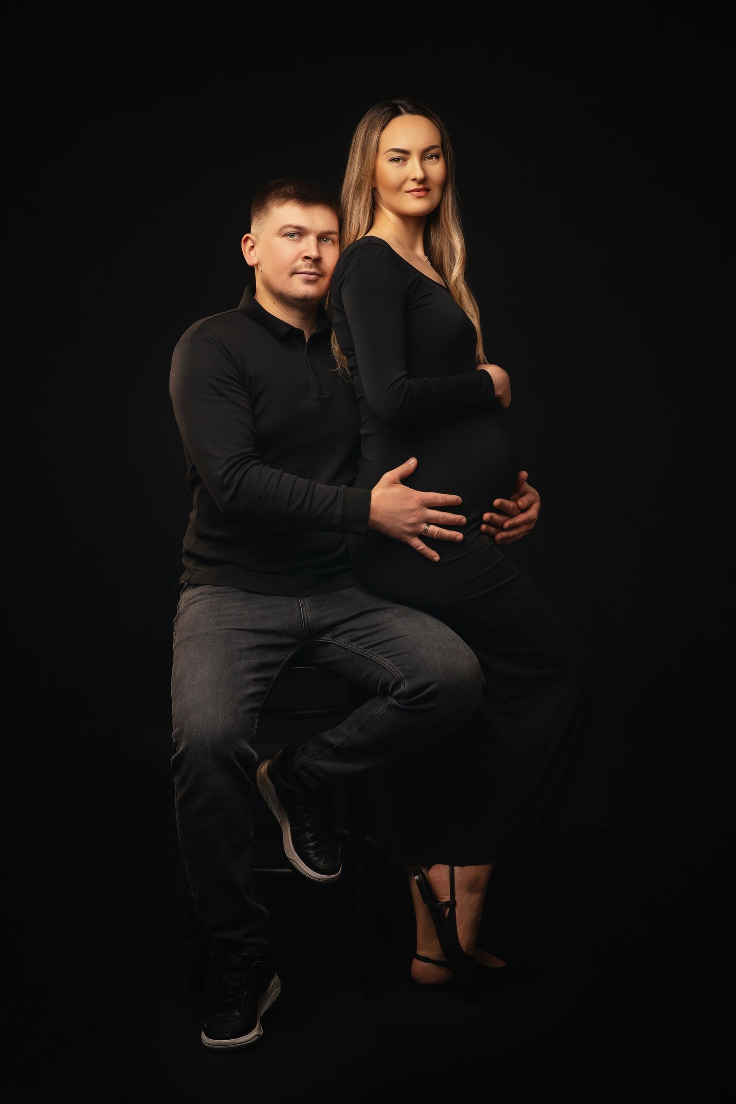 A couple poses together in a dark studio setting for a maternity photo, both wearing black against a black background.