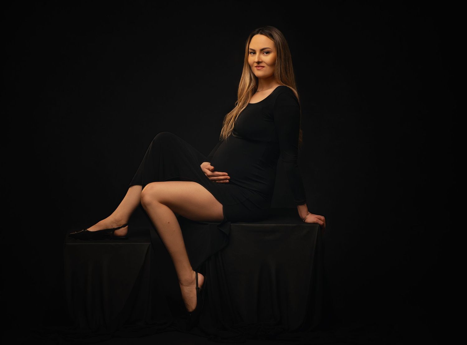 A figure in a black dress poses elegantly on a dark pedestal against a dramatic black background.
