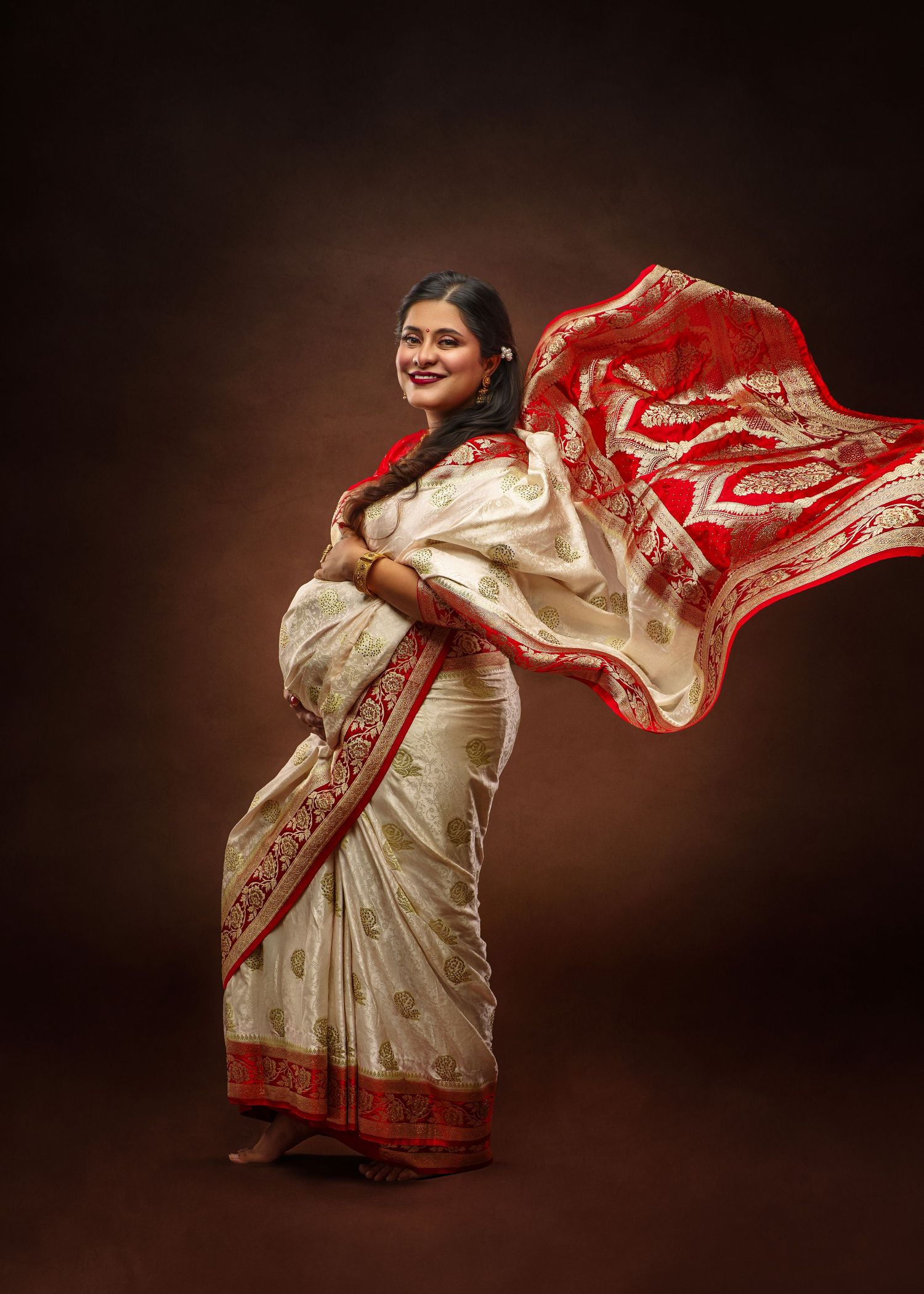 Traditional Indian cream and red silk saree with flowing fabric against dark brown backdrop in elegant portrait pose.