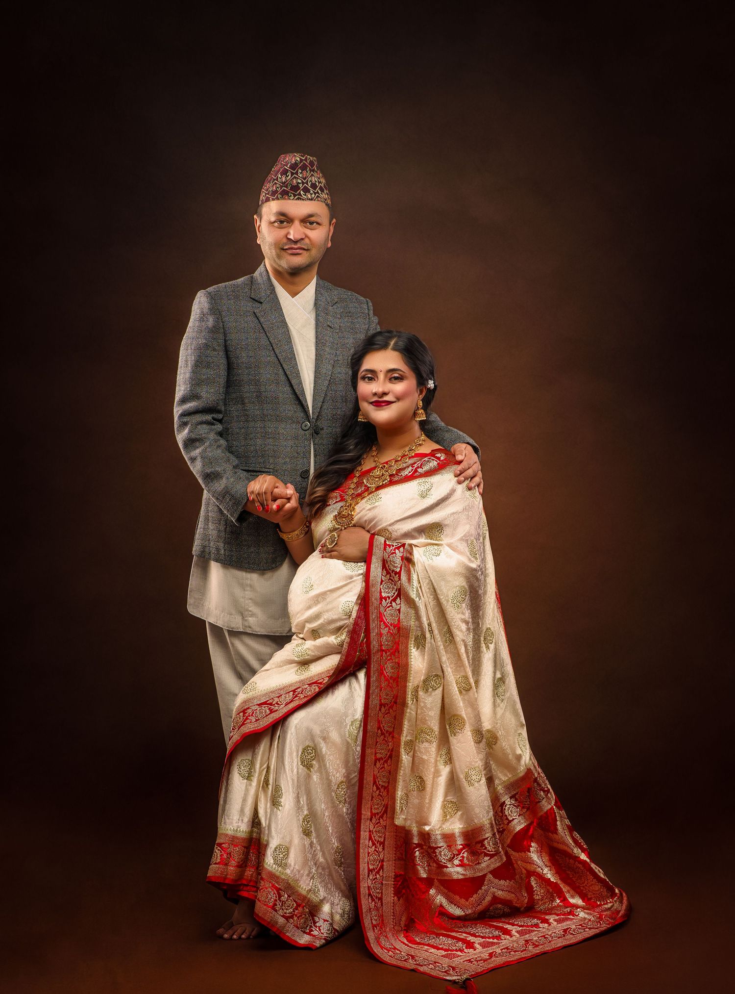 A couple poses in traditional Nepalese attire with the woman wearing an elegant white and red saree in a studio setting.