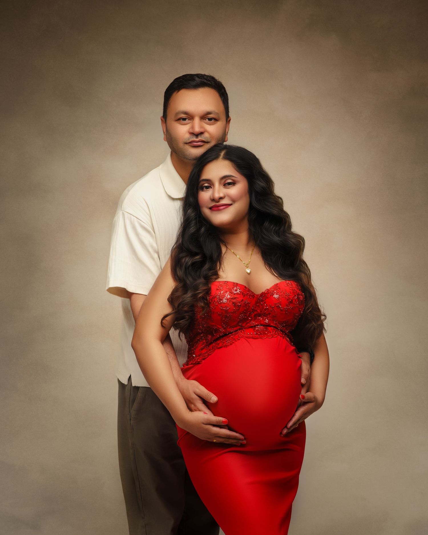 A couple poses together in a maternity photo, with the pregnant woman wearing an elegant red formal dress against a brown background.