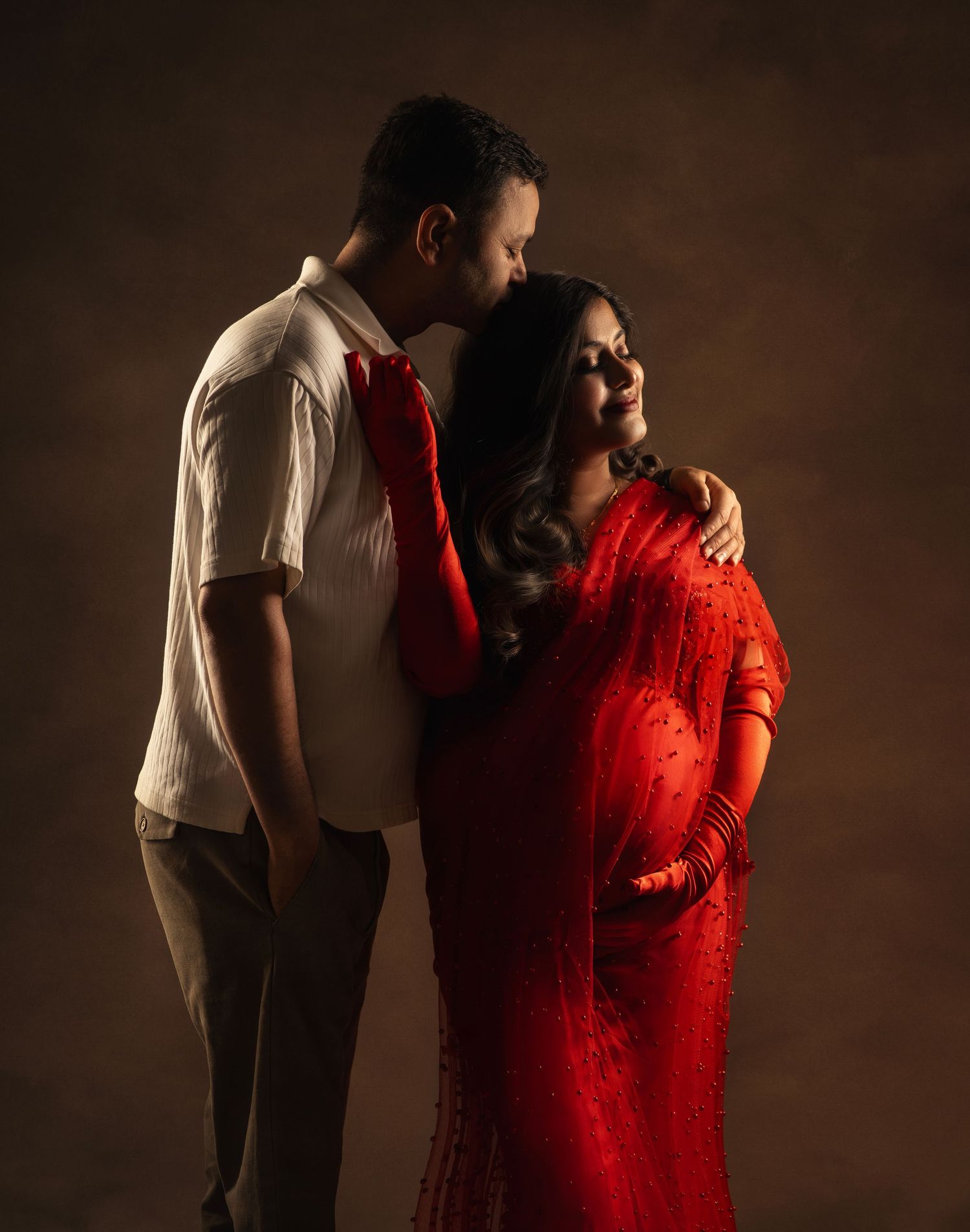 A couple embraces in a romantic studio portrait with moody lighting, the woman wearing a vibrant red traditional dress.