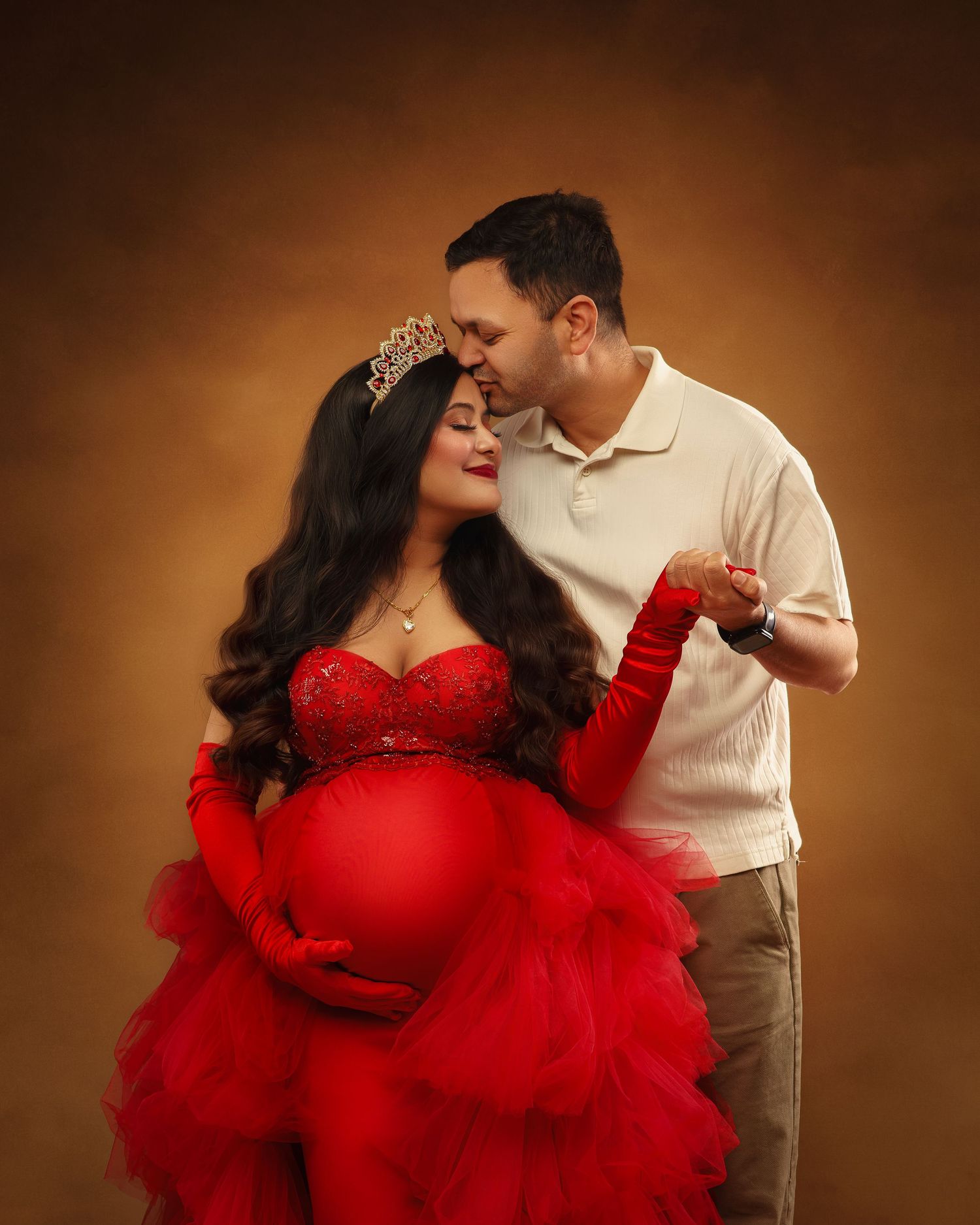 Romantic maternity photo featuring a woman in a glamorous red tulle gown with a tiara posing with her partner in warm lighting.
