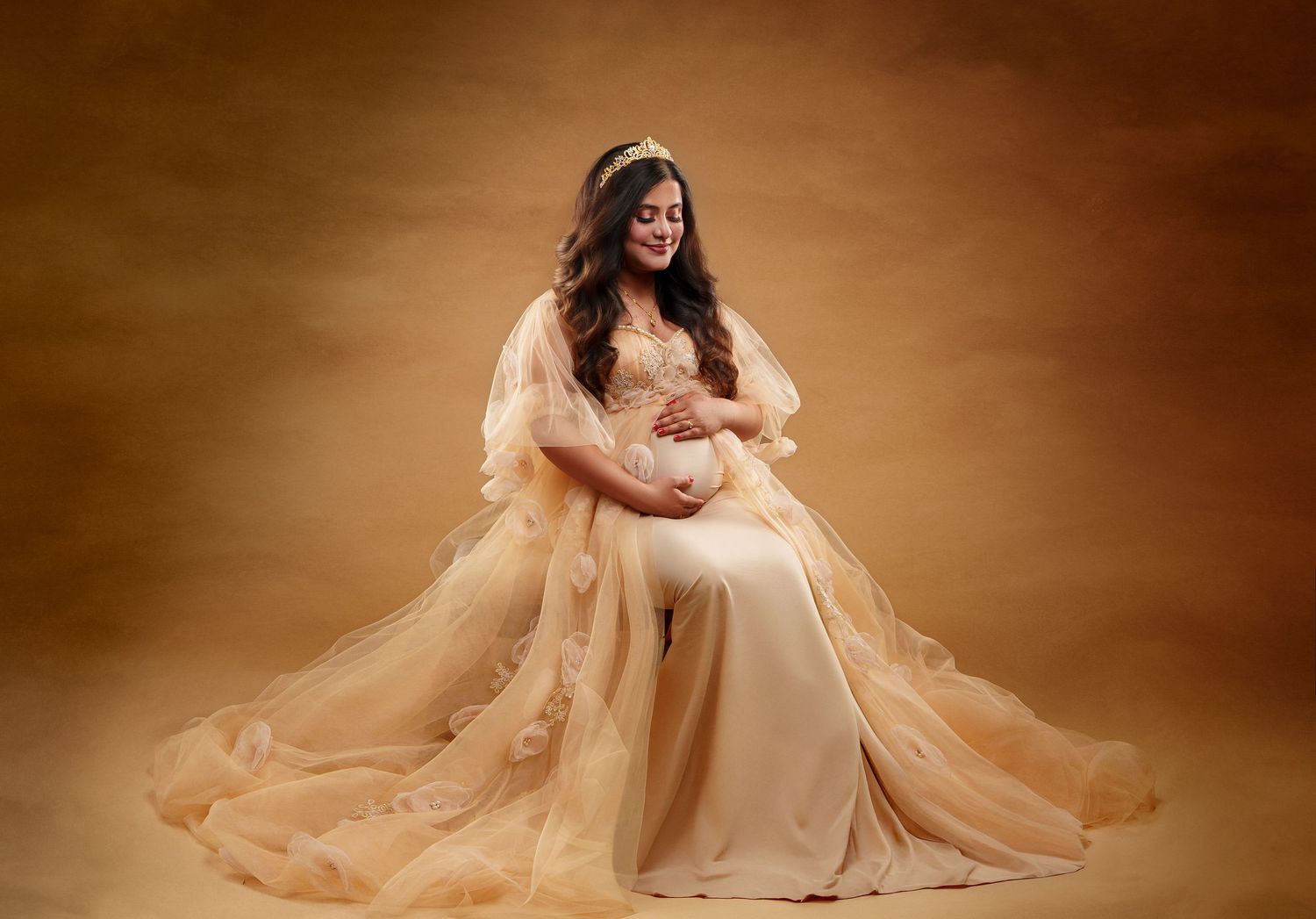 Elegant maternity portrait featuring a flowing champagne-colored gown against a warm brown studio backdrop.
