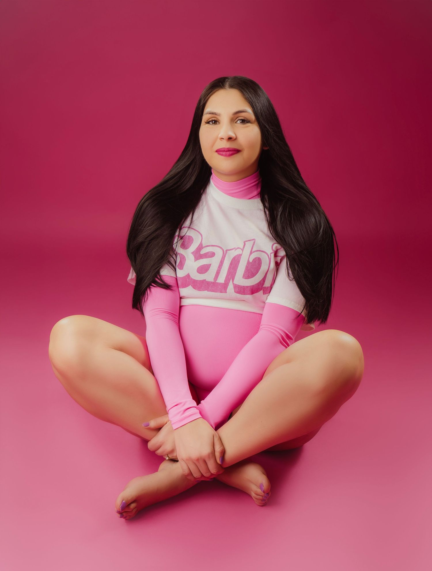 Someone poses in pink athletic wear with long dark hair against a pink background in a cross-legged seated position.