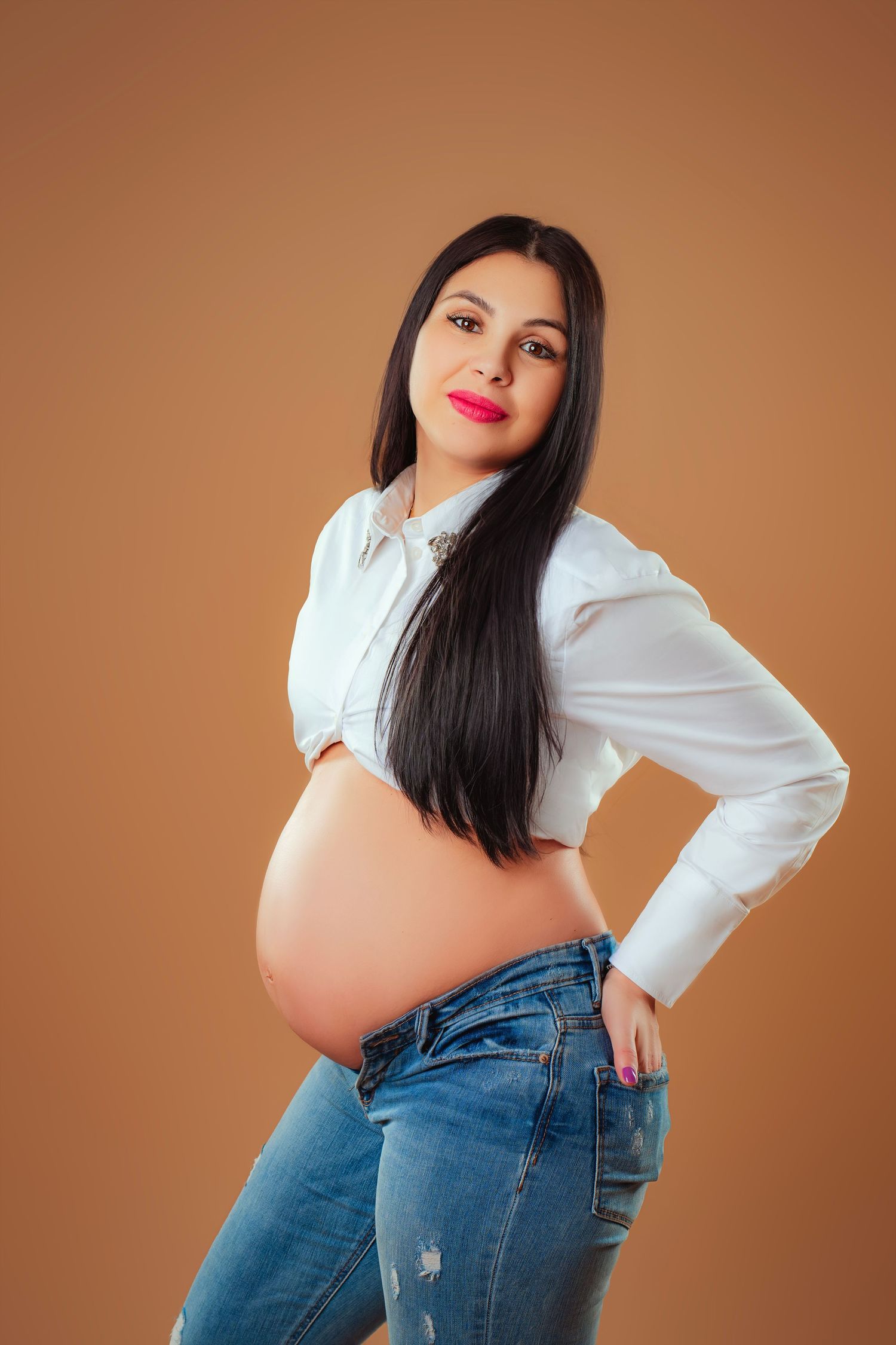 A pregnant woman in a white button-up shirt and blue jeans poses against a brown backdrop.