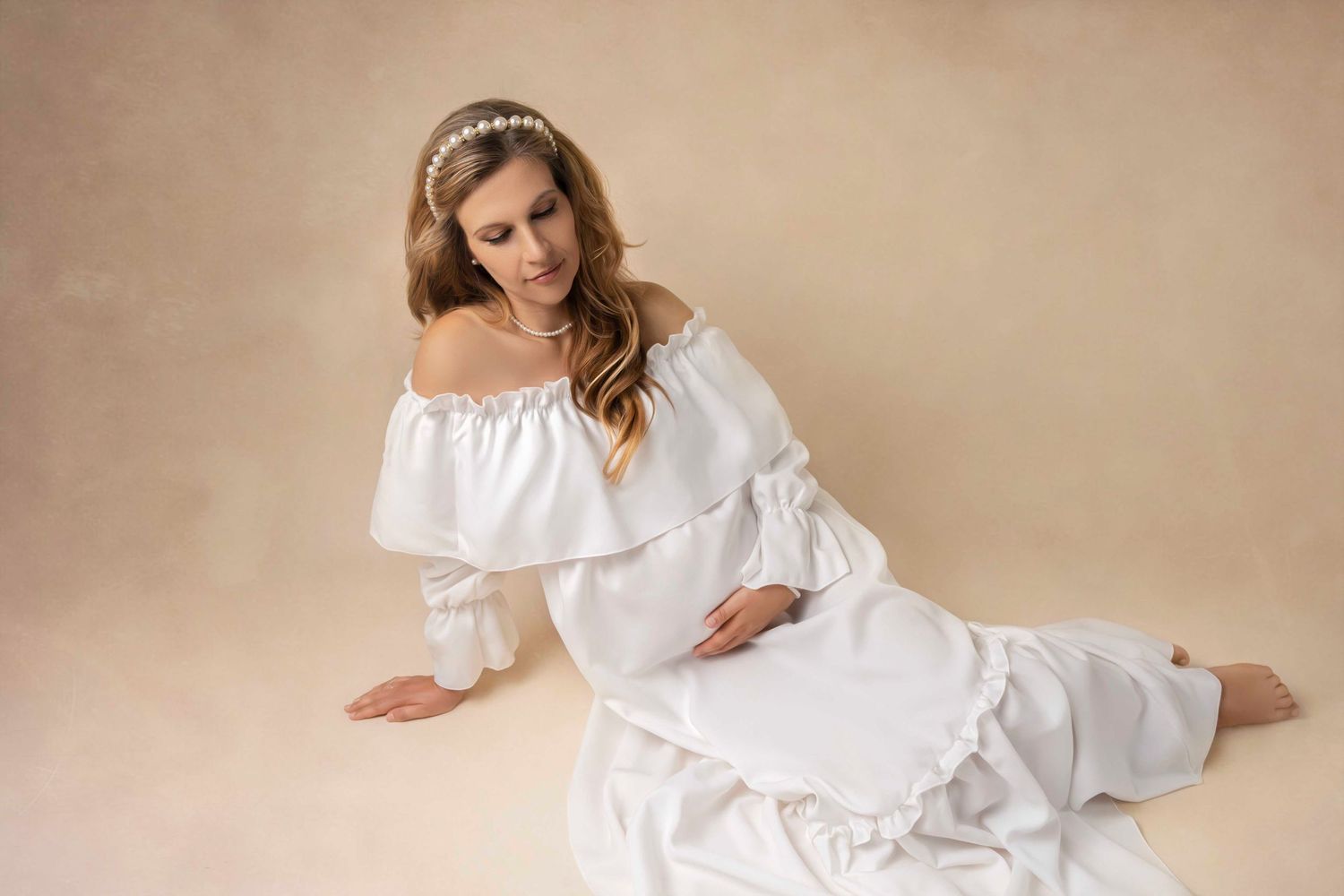 Woman in flowy white off-shoulder dress lying on neutral beige background in romantic vintage inspired portrait.
