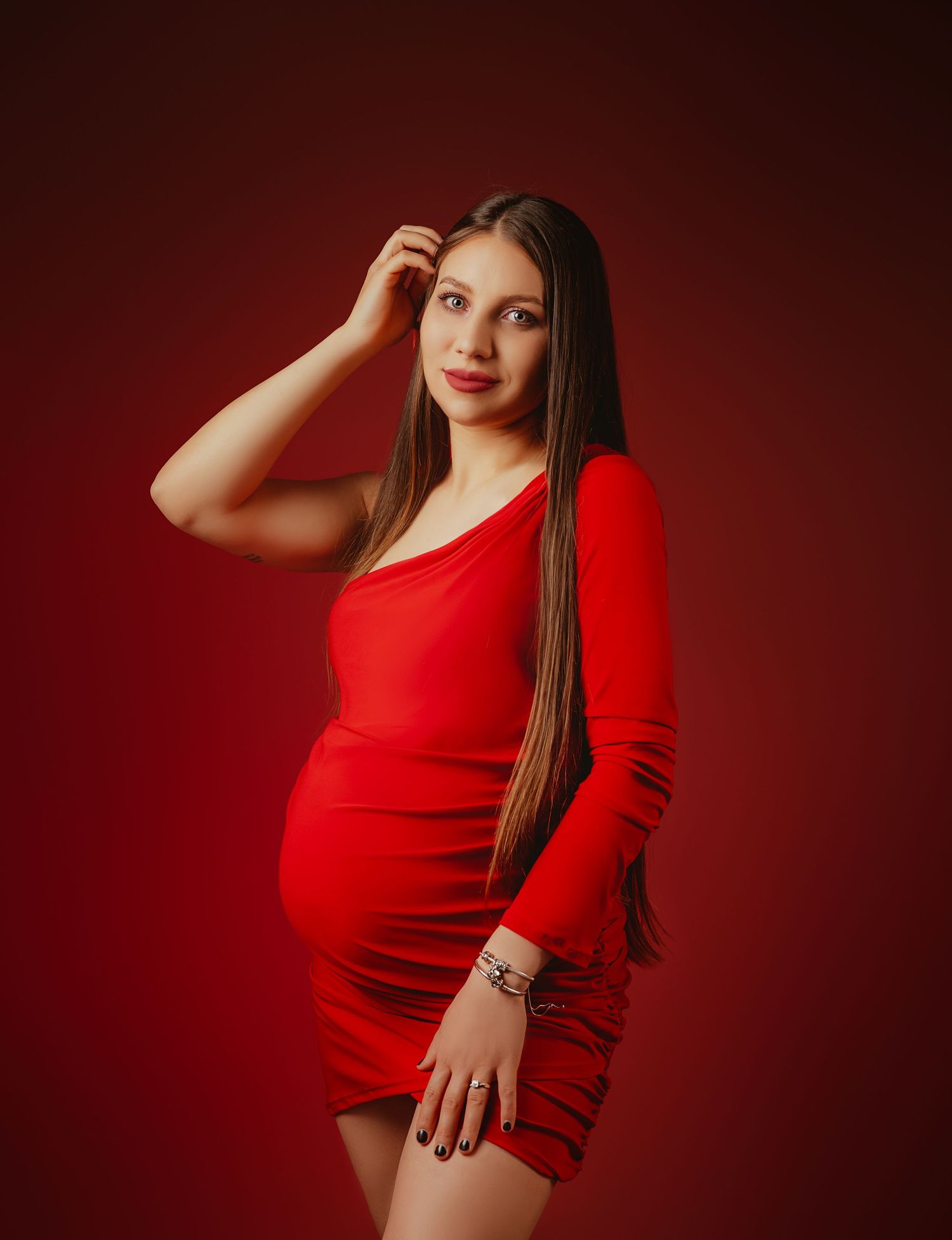 Maternity portrait against deep red background featuring long straight hair and fitted red dress.