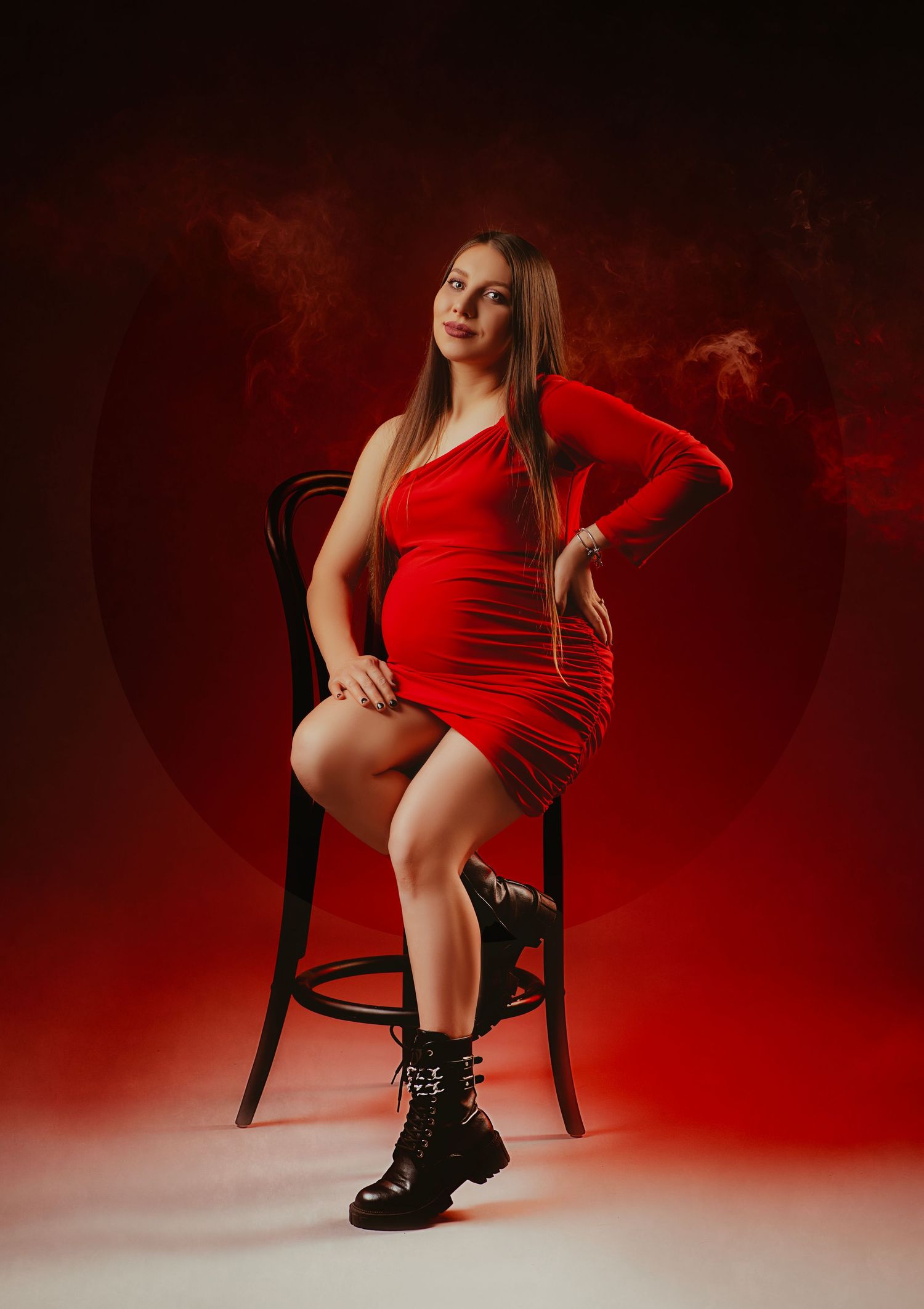Artistic portrait against red background featuring person in red dress seated on dark wooden chair wearing combat boots.
