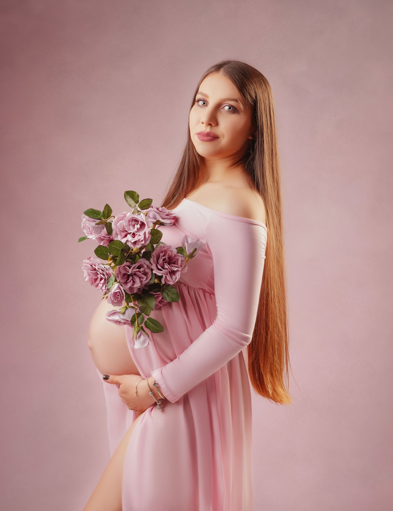 A dreamy maternity portrait with flowing pink dress and roses against a rosy backdrop.