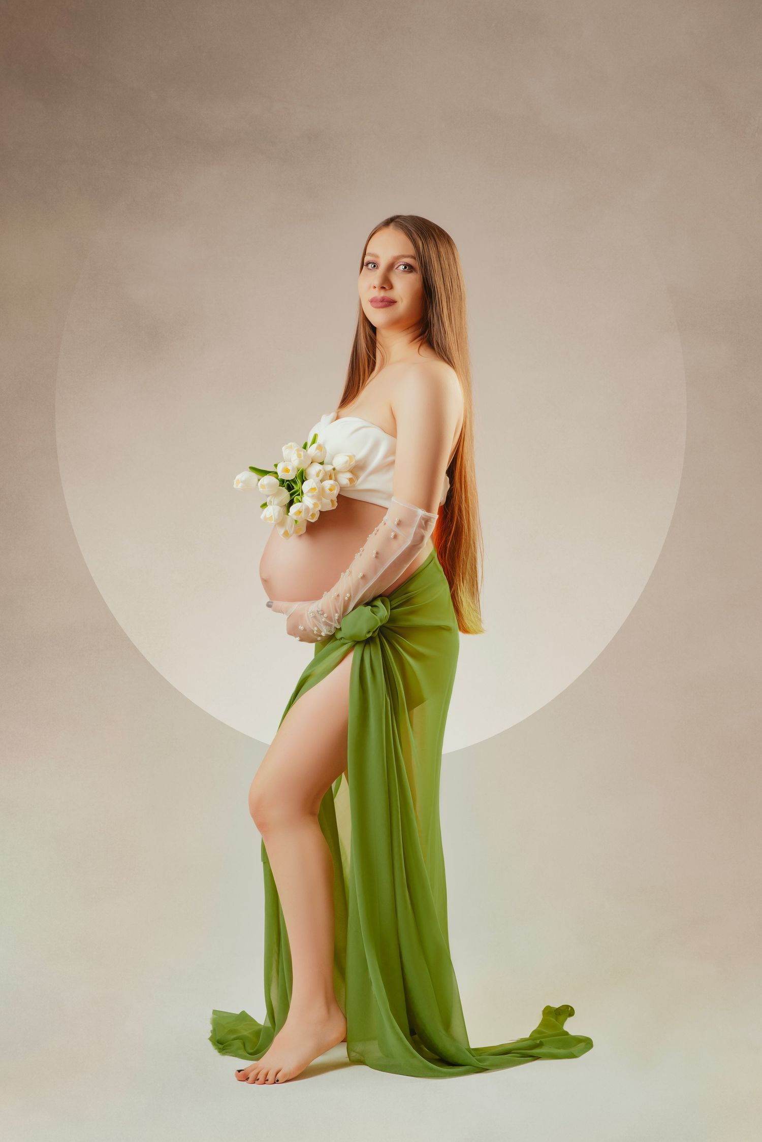 Expecting mother in green draped fabric holds white flowers during elegant maternity photoshoot against neutral backdrop.