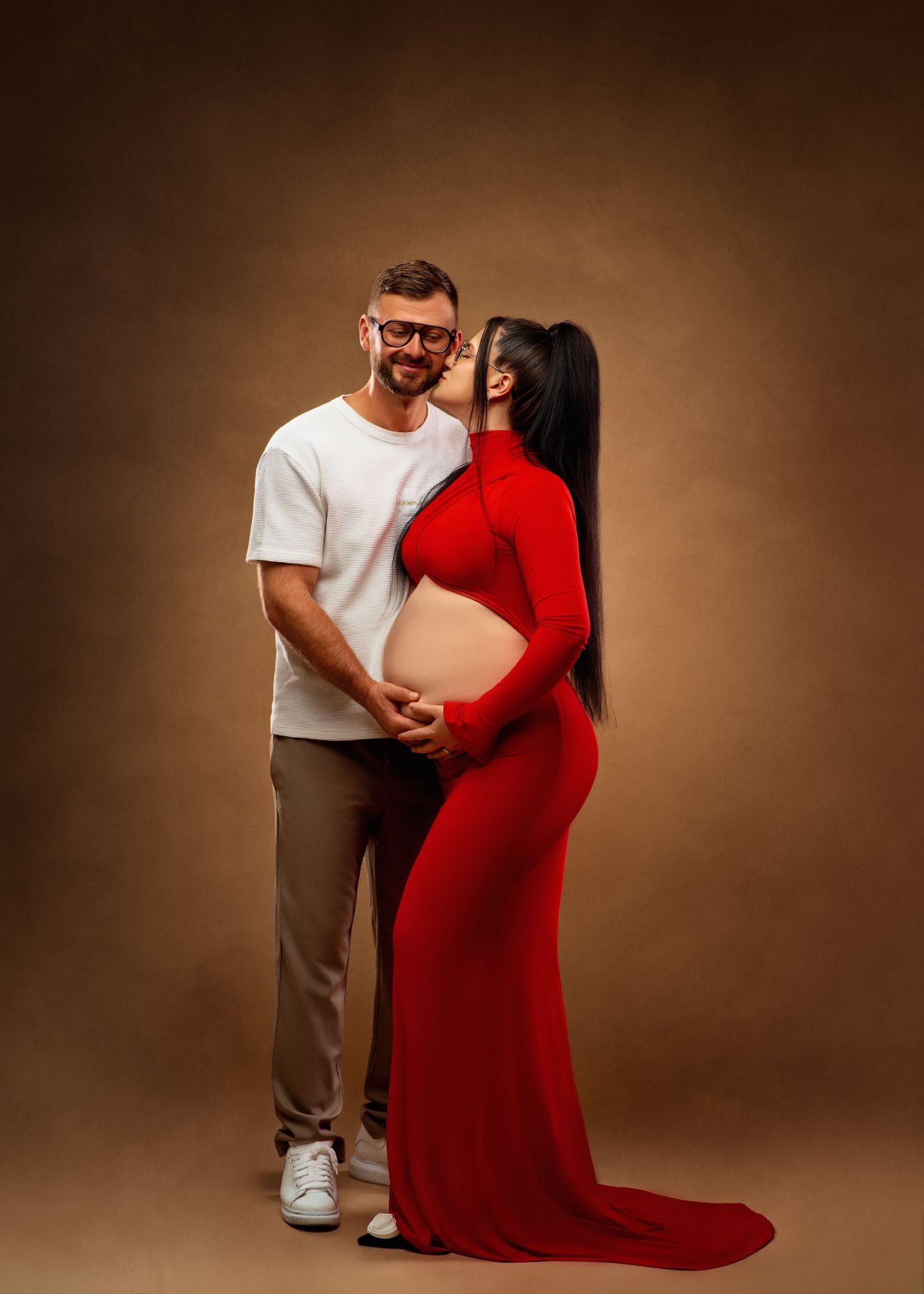 A couple shares an intimate moment during a maternity photo shoot against a brown backdrop, with her wearing a striking red gown.