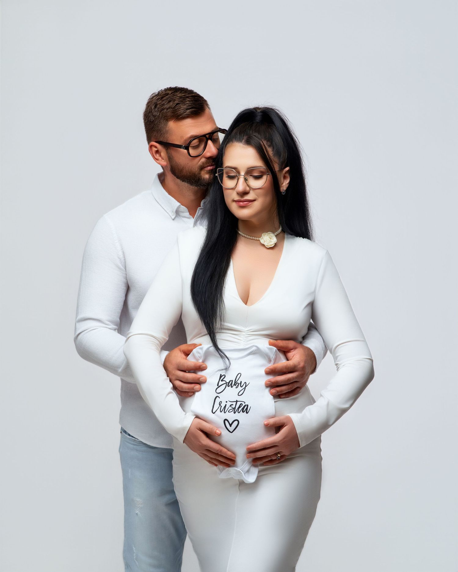 A couple in matching white outfits and glasses embrace while showcasing a pregnancy reveal against a light grey background.