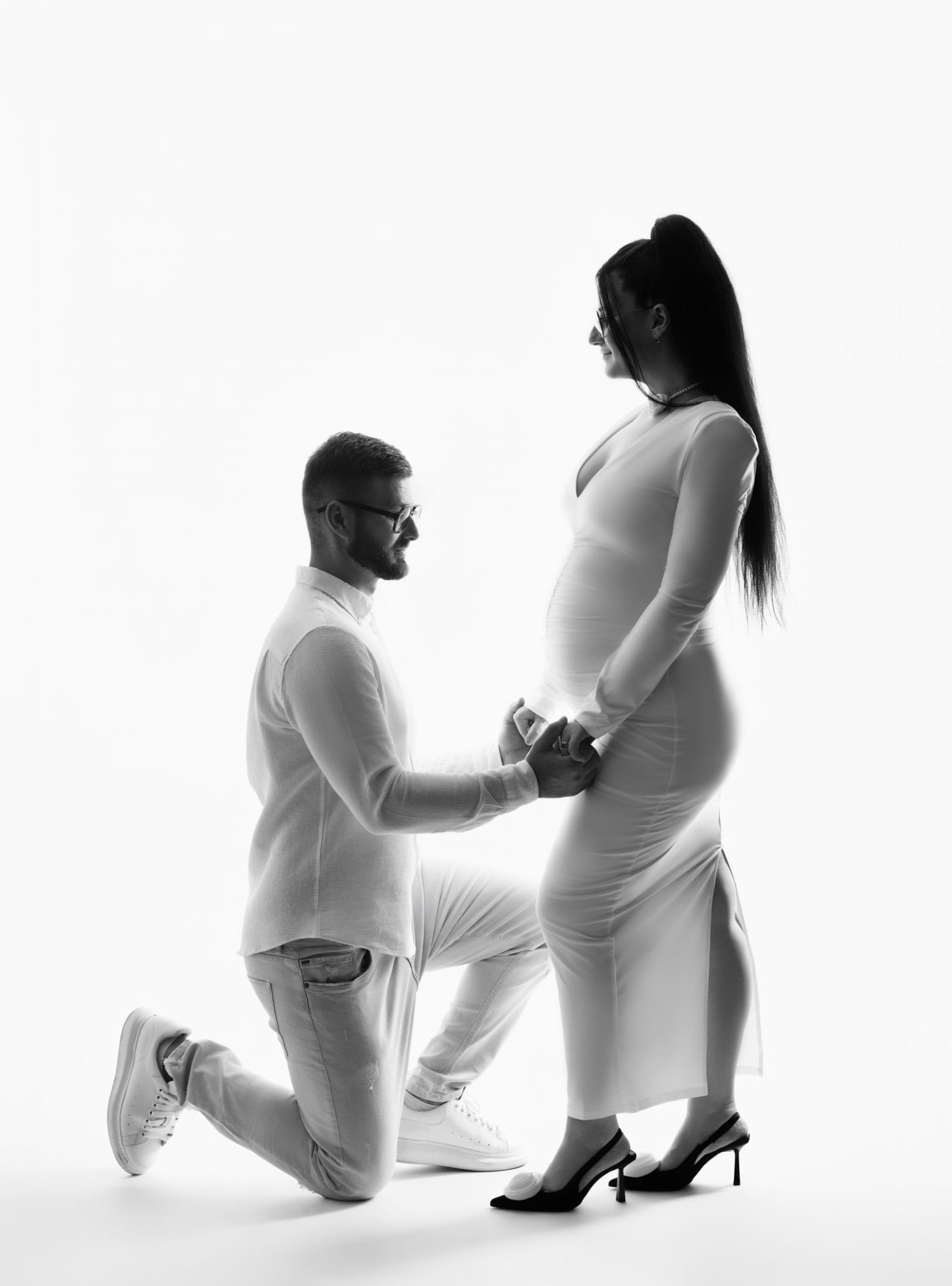 A couple shares an intimate moment in a minimalist white studio setting, with one person kneeling before an expectant mother.