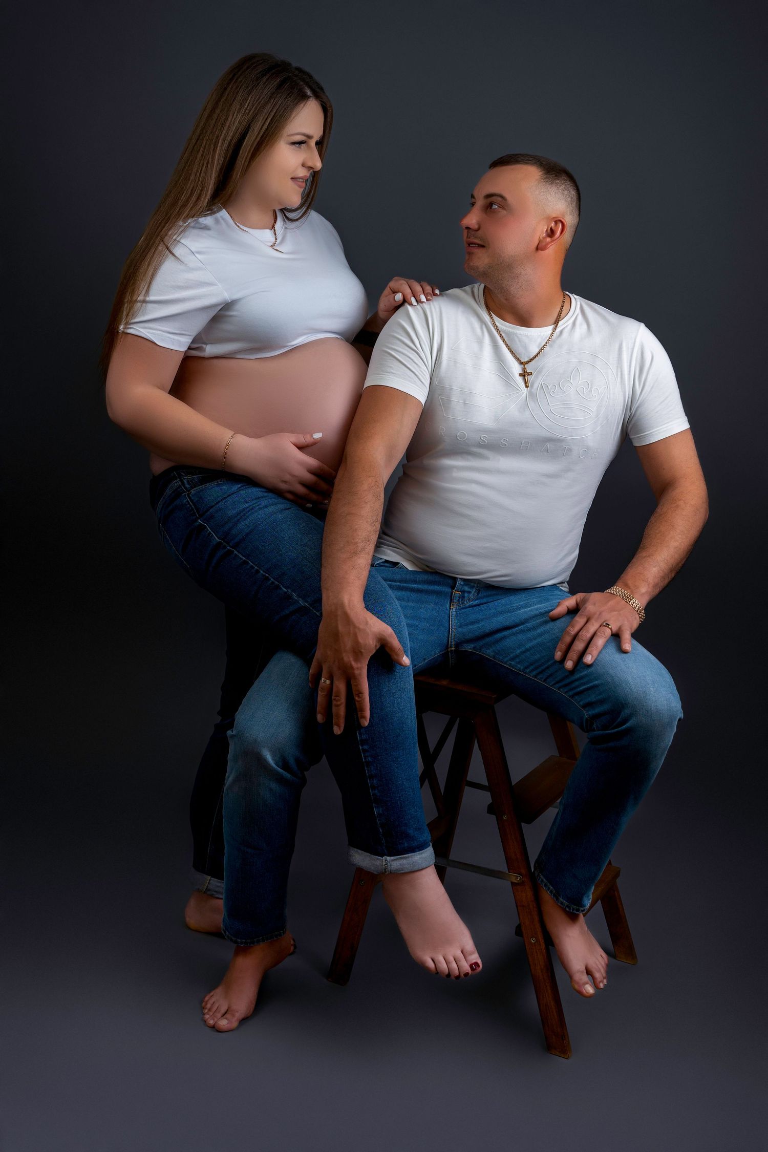 A couple in matching white shirts and jeans poses for a pregnancy photoshoot against a dark background.