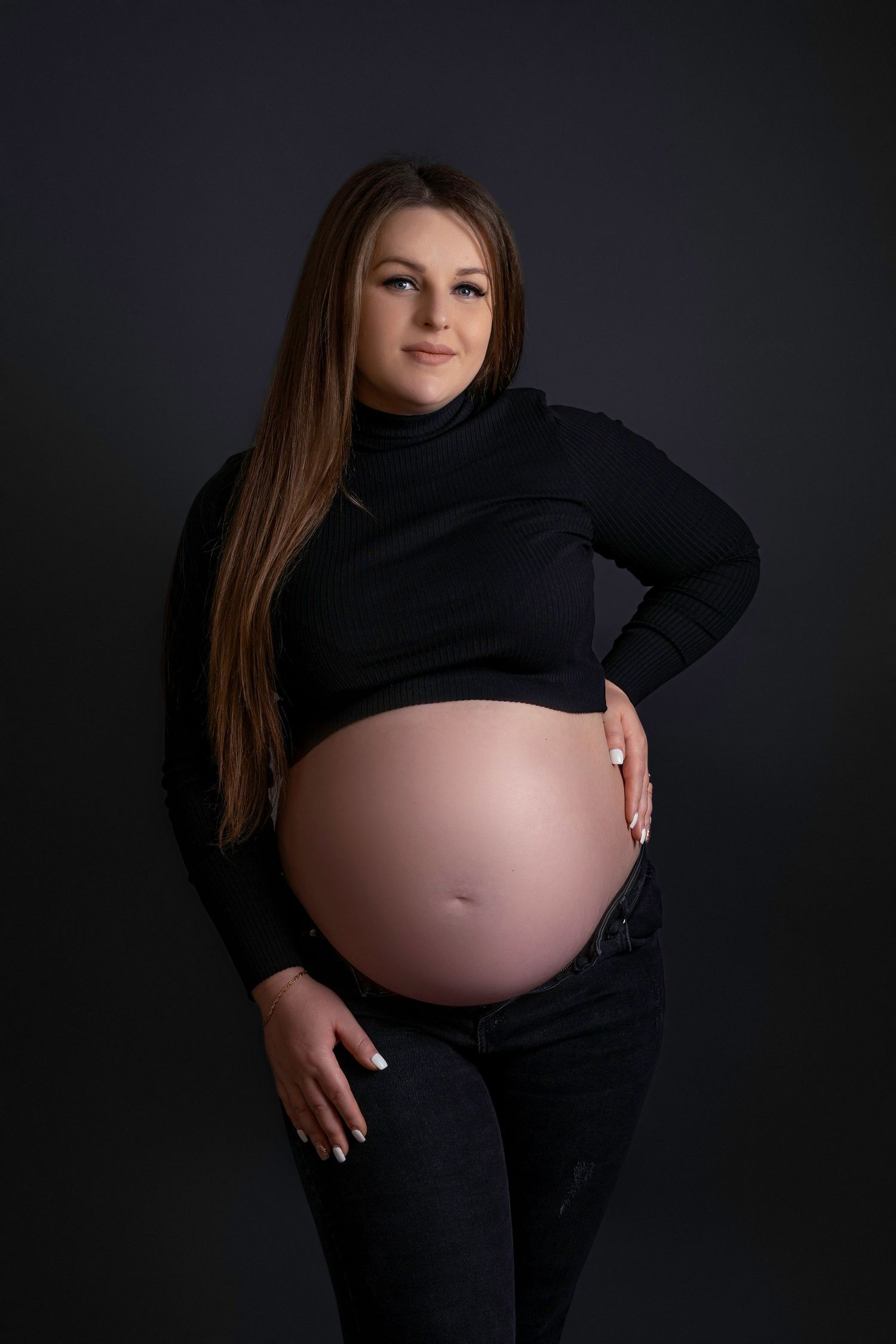 Maternity portrait of an expectant mother in black clothing against dark background showing her pregnant belly.