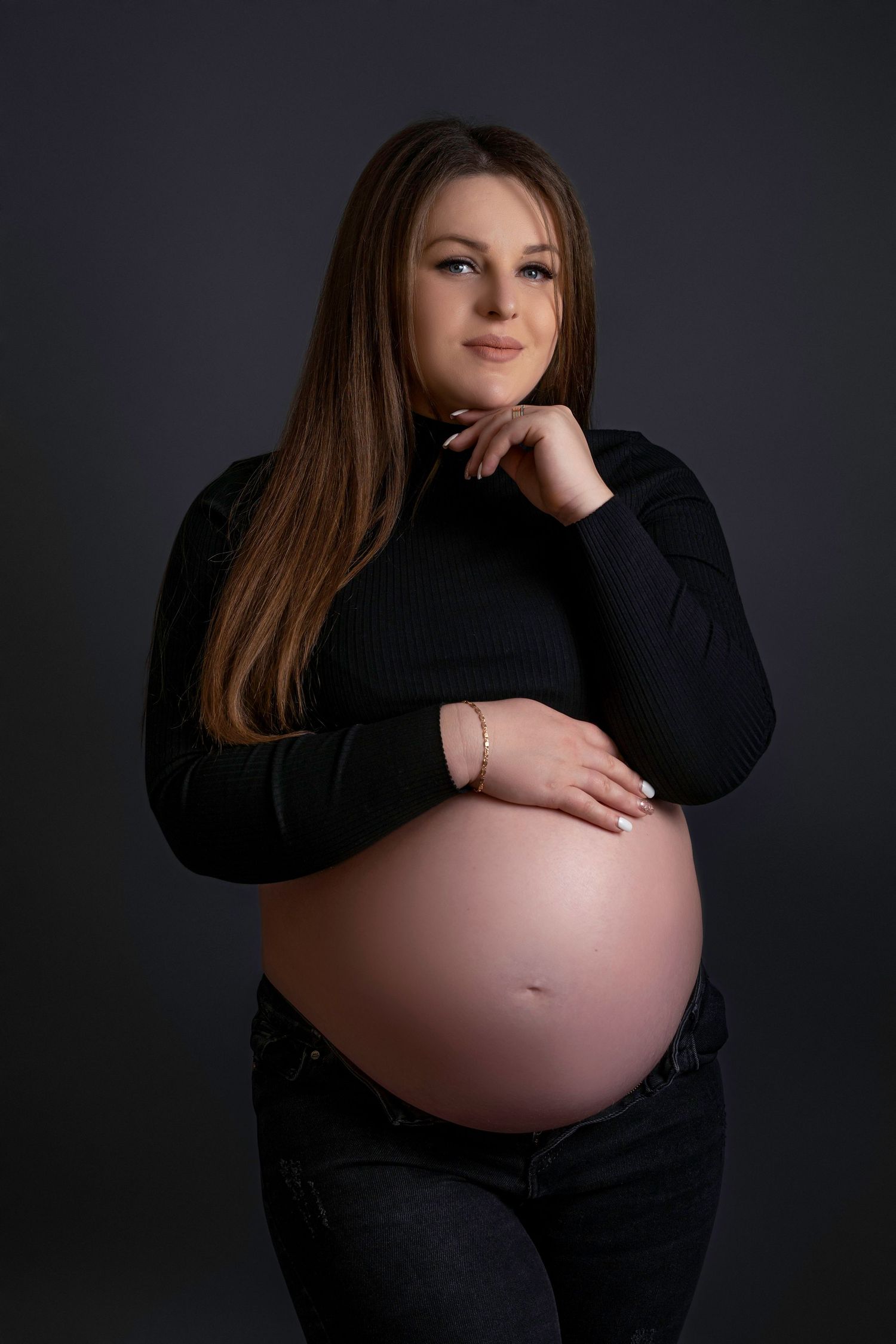 A pregnant woman in a black sweater poses against a dark background for a maternity photo.