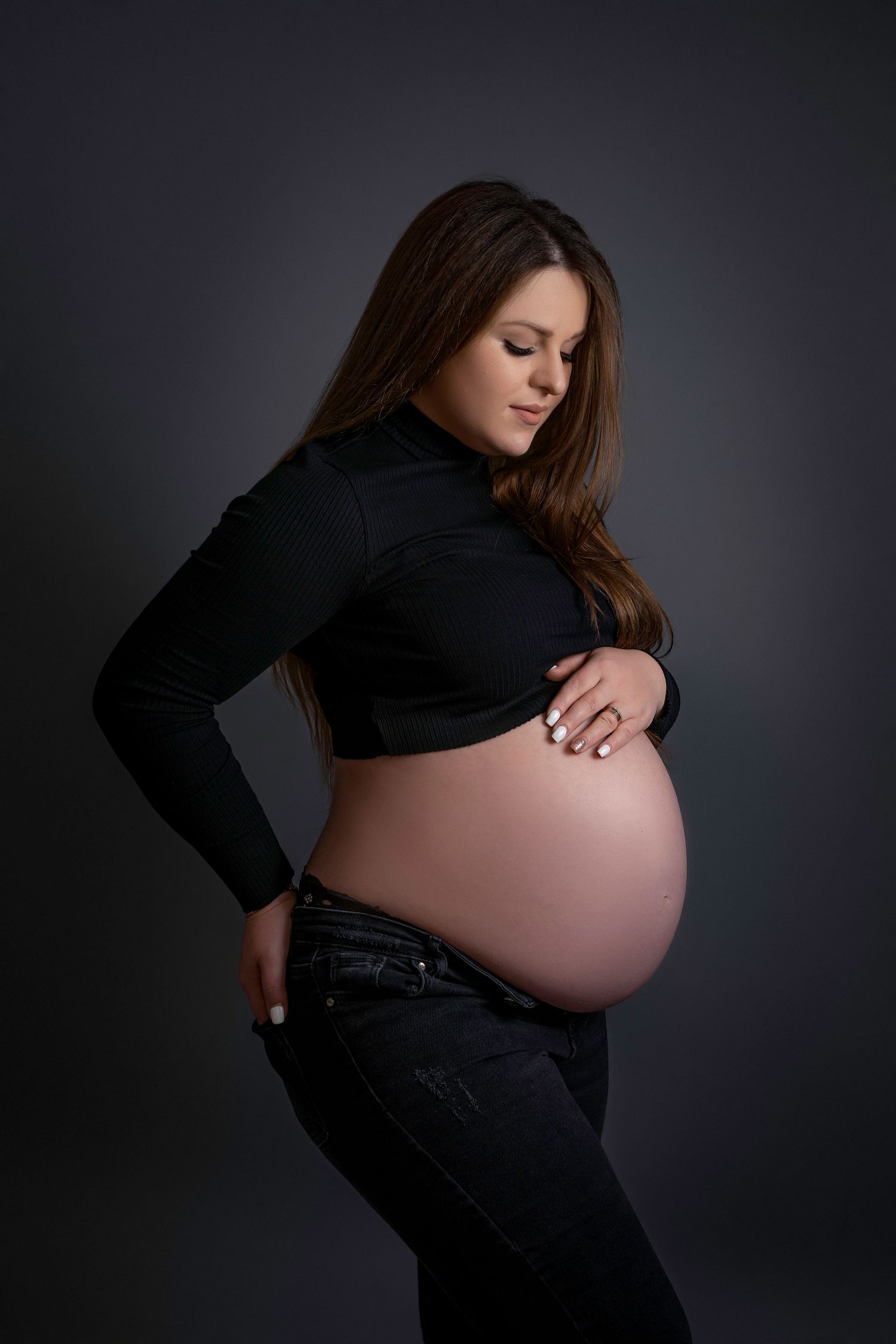 Elegant maternity portrait showing a pregnant woman in black turtleneck against dark background, highlighting her baby bump.