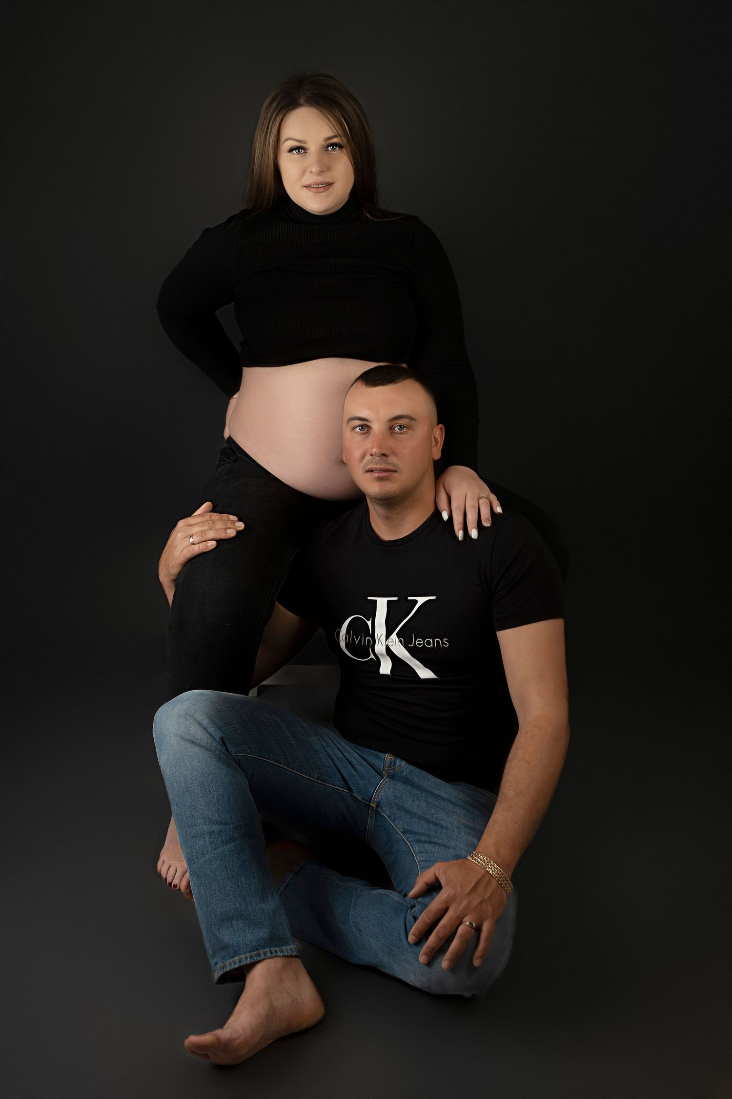 A couple poses for a dramatic maternity photo against a dark background wearing black clothing.
