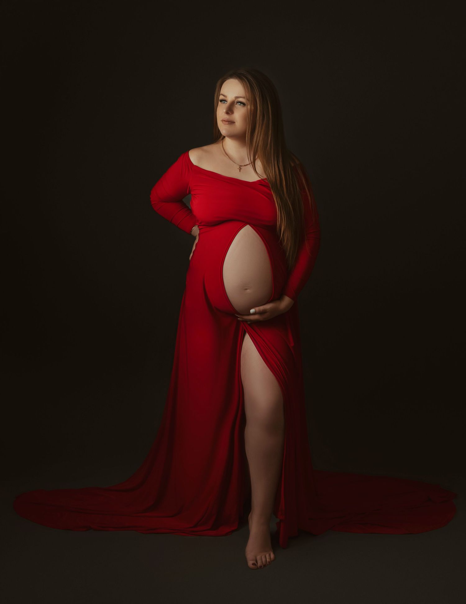 Maternity portrait featuring a long red gown against a dark background in dramatic lighting.