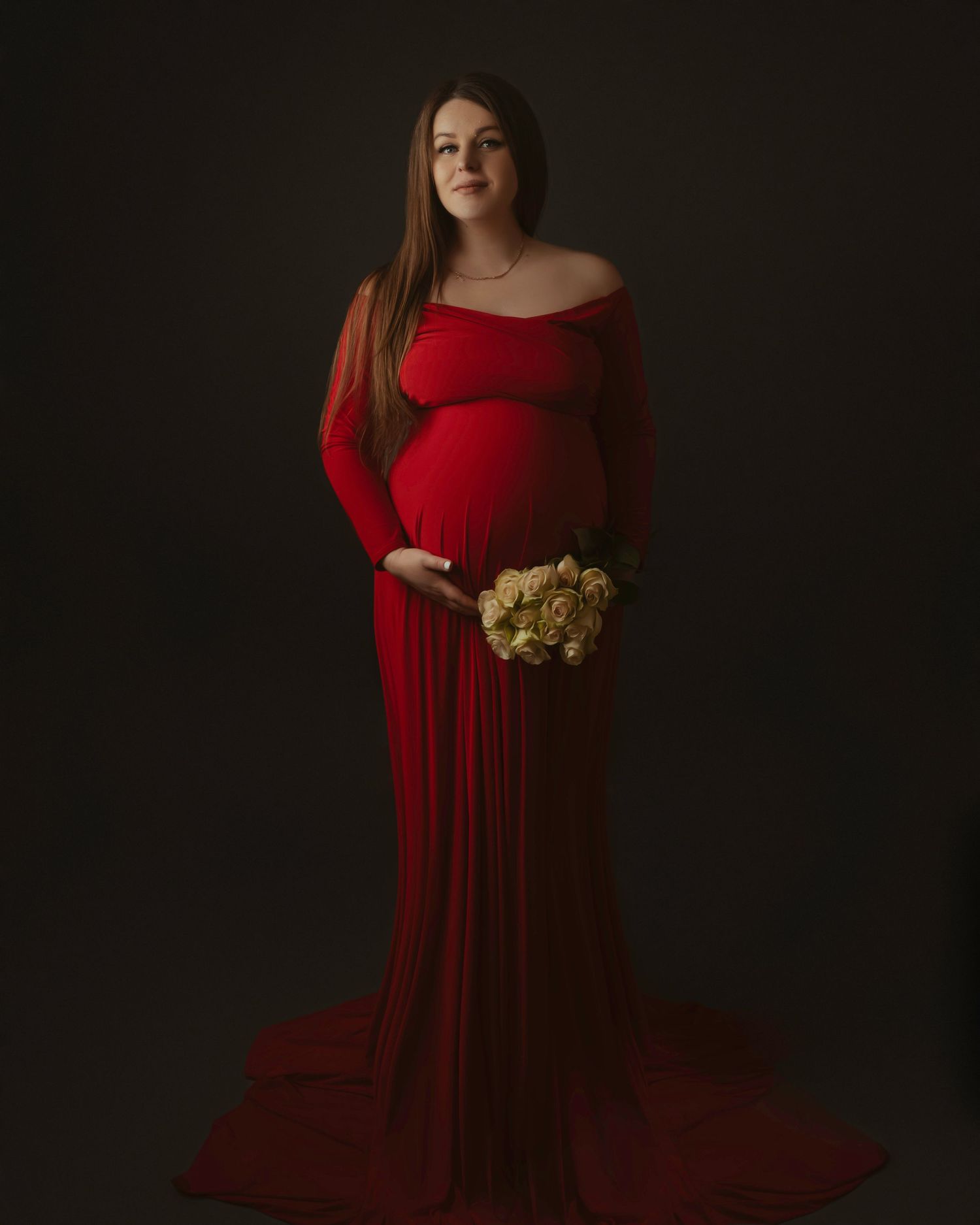 Maternity portrait in dramatic lighting features an expectant mother in a flowing red gown holding white flowers.