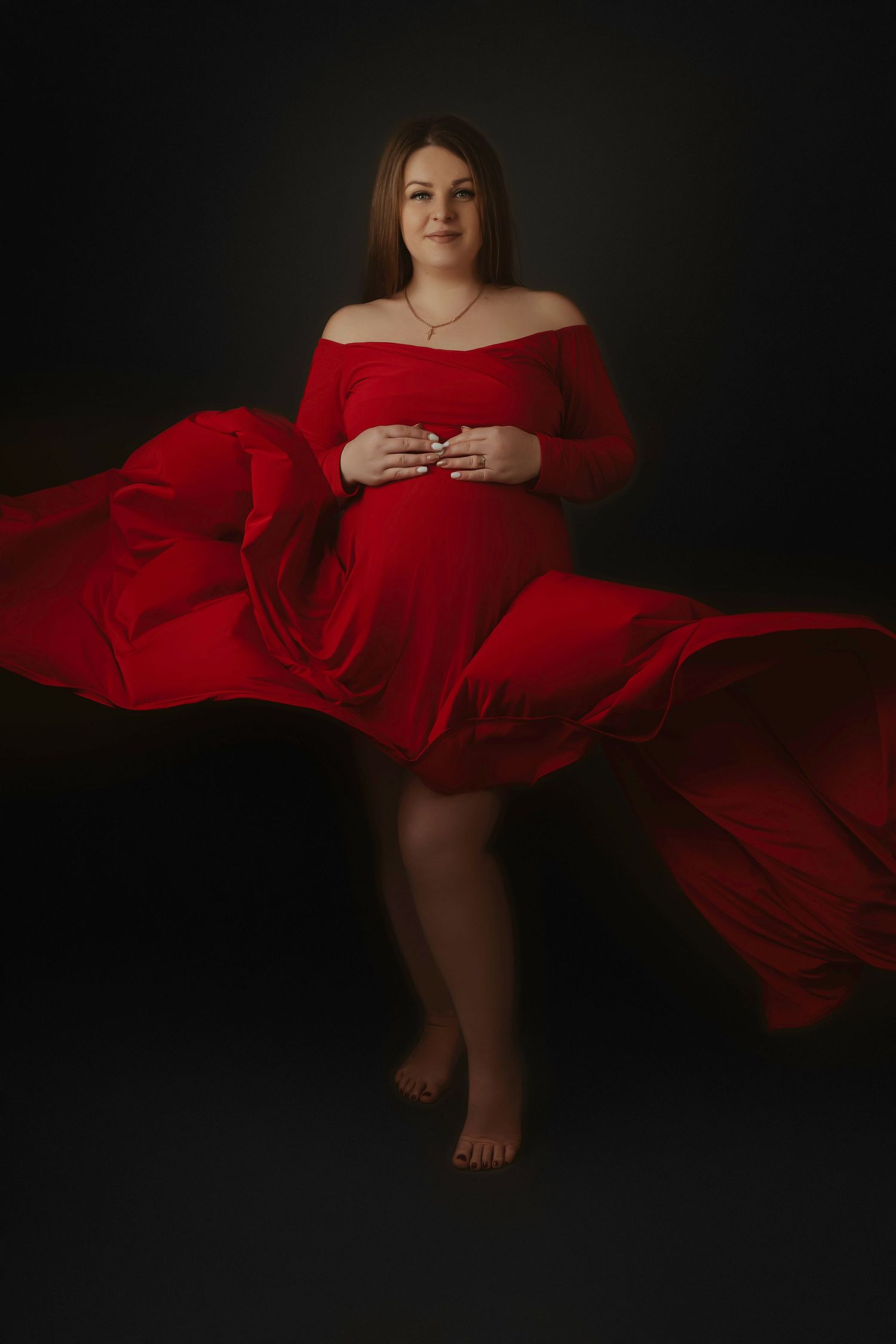 Maternity portrait in flowing red gown against dark background with dramatic fabric movement and off-shoulder design.
