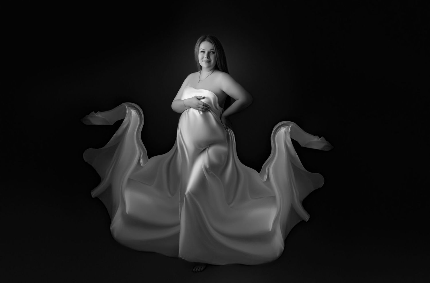 Black and white maternity portrait with flowing white fabric creating dramatic movement against dark background.