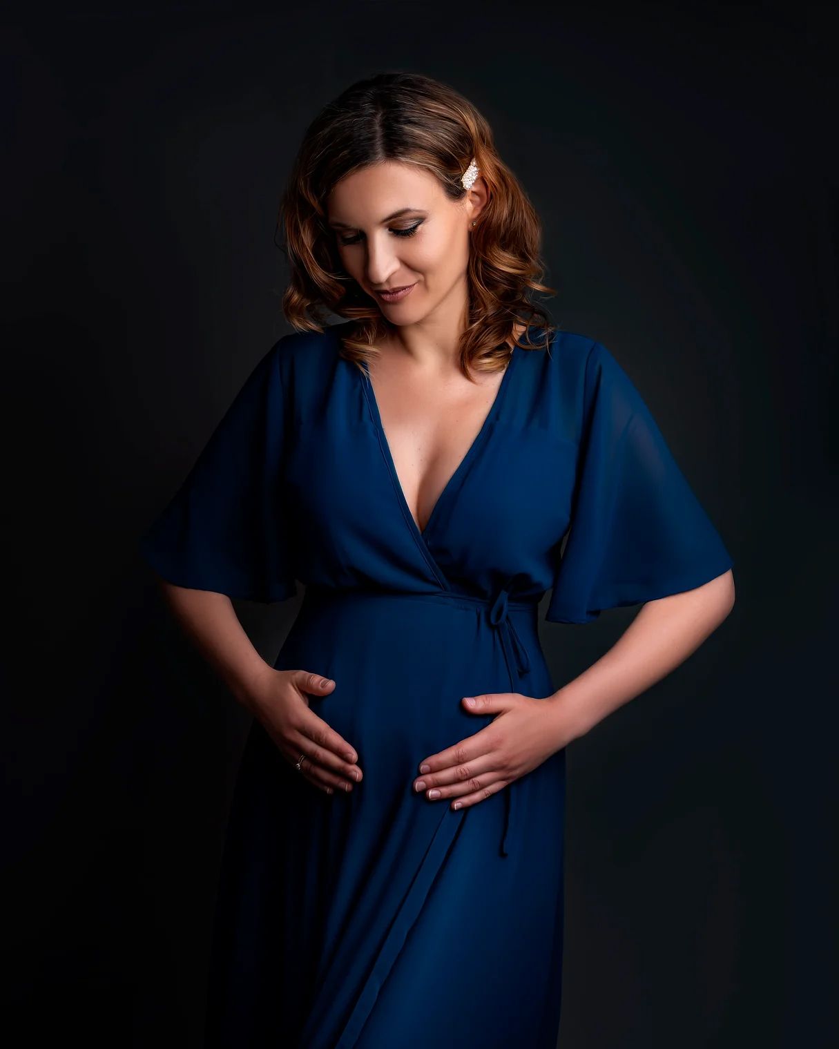 Expectant woman in elegant navy blue wrap dress poses against dark backdrop during maternity photoshoot.