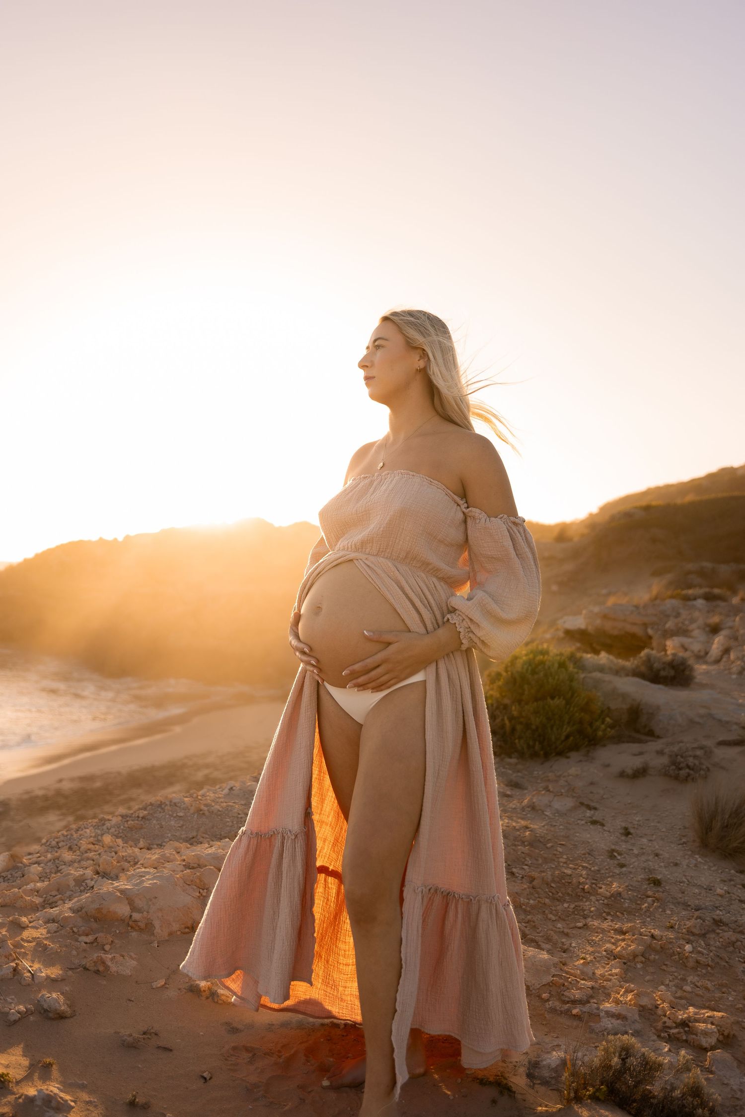 Pregnant woman in flowing dress stands on cliff during sunset.