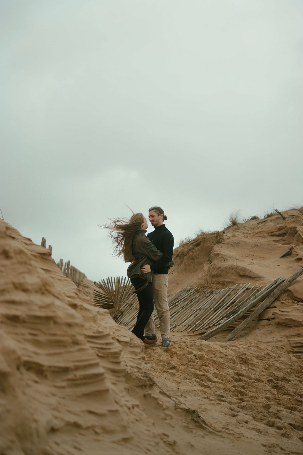 A couple stands atop coastal sand dunes with beach fencing visible.