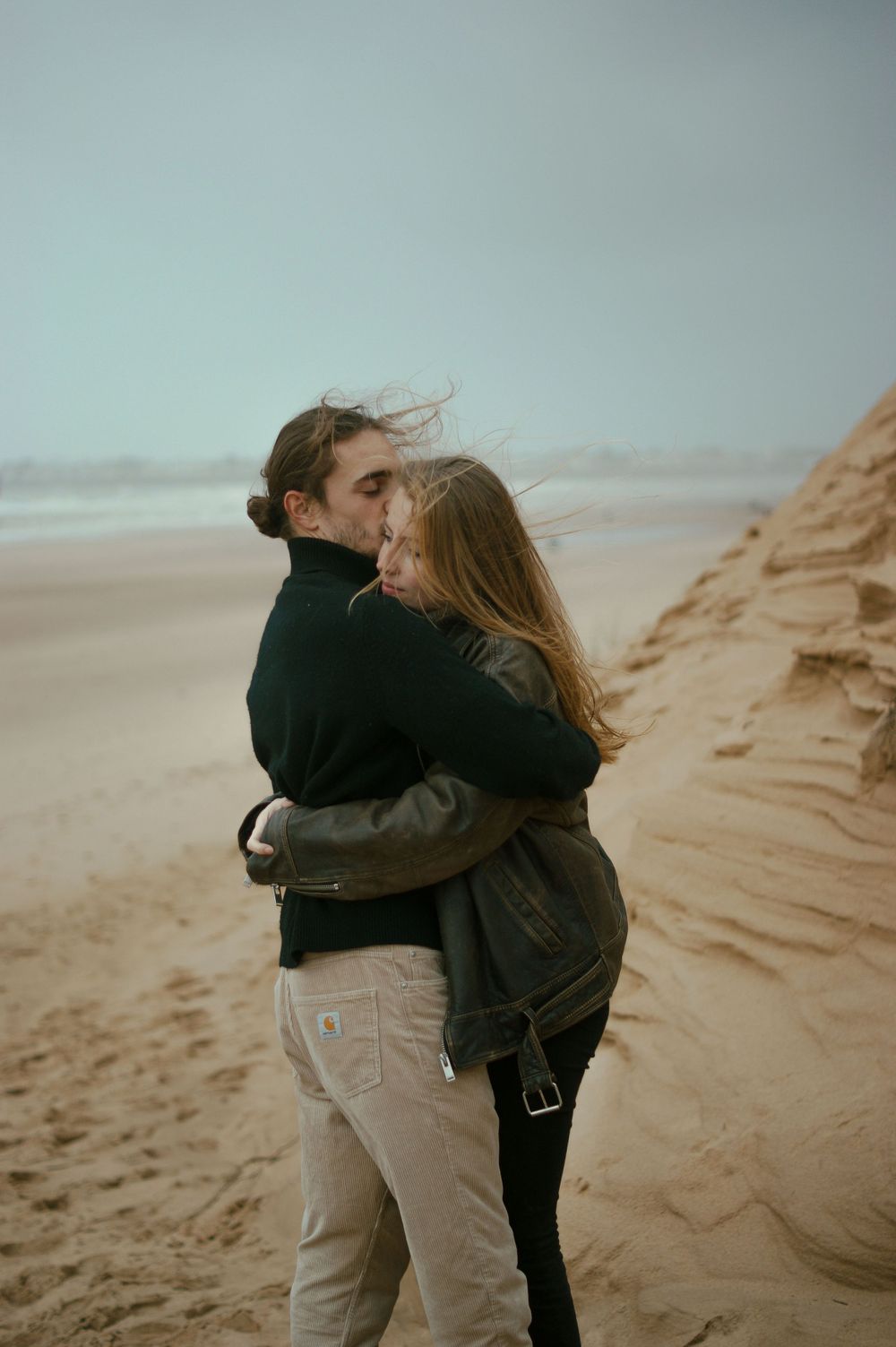 A romantic embrace on windy sand dunes with the ocean in the background.