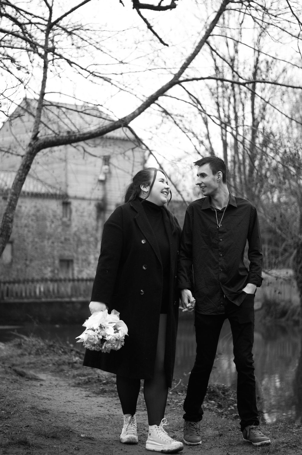 A couple in dark clothing stands by a pond with bare trees and buildings in the background.