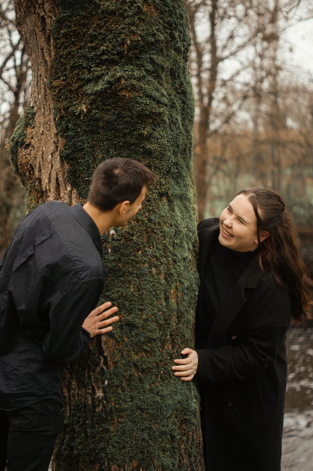 A playful couple shares a moment around a moss-covered tree trunk in a forest setting.