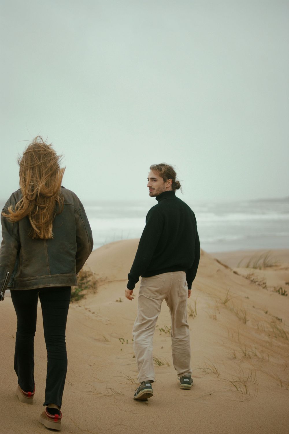 Two people walking on windswept coastal sand dunes under an overcast sky.