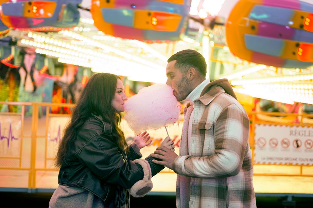 Couple sharing a playful moment at a carnival with illuminated rides in background.
